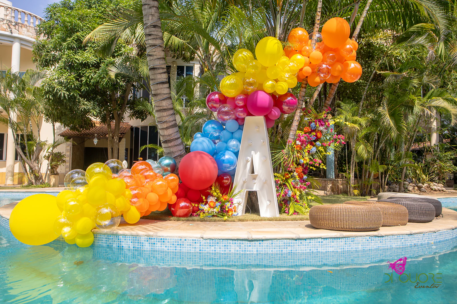 A grande letra A com balões amarelos, laranja, azuis e cor-de-rosa e ramos de flores tropicais, numa plataforma rodeada pela água da piscina