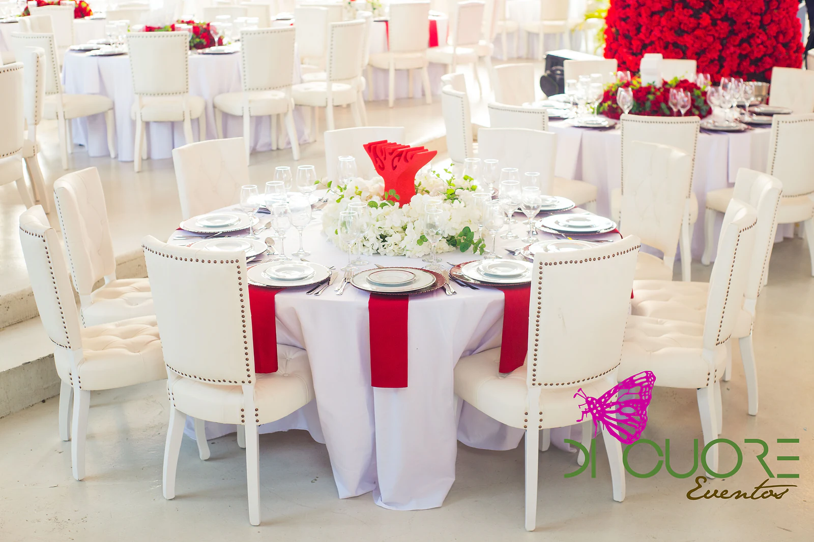 Round table in white linen with red napkins, red chargers, a centrepiece of white flowers and tufted white chairs