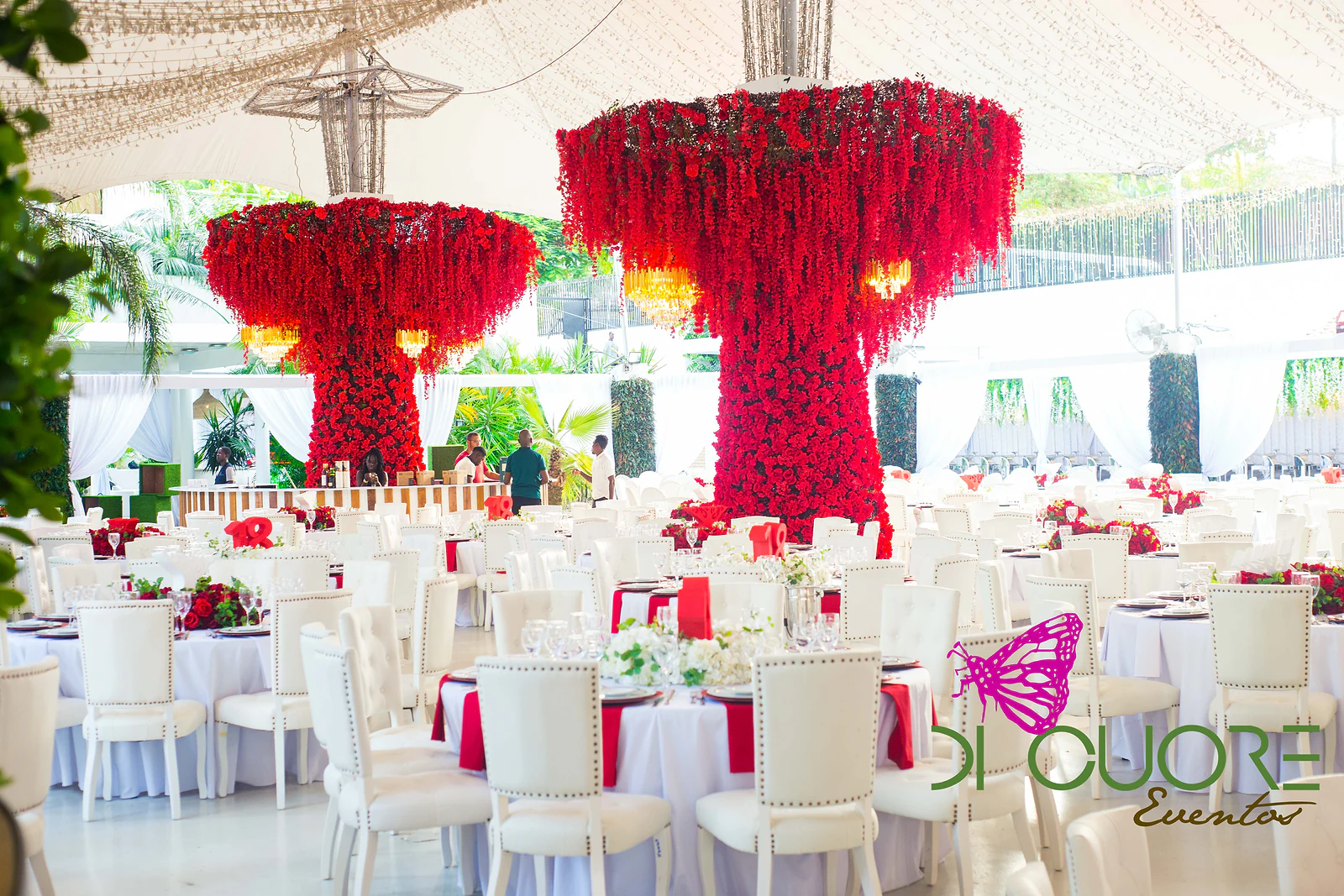 Two hanging chandeliers covered in red flowers above round tables in white linen with crimson napkins and tufted white chairs