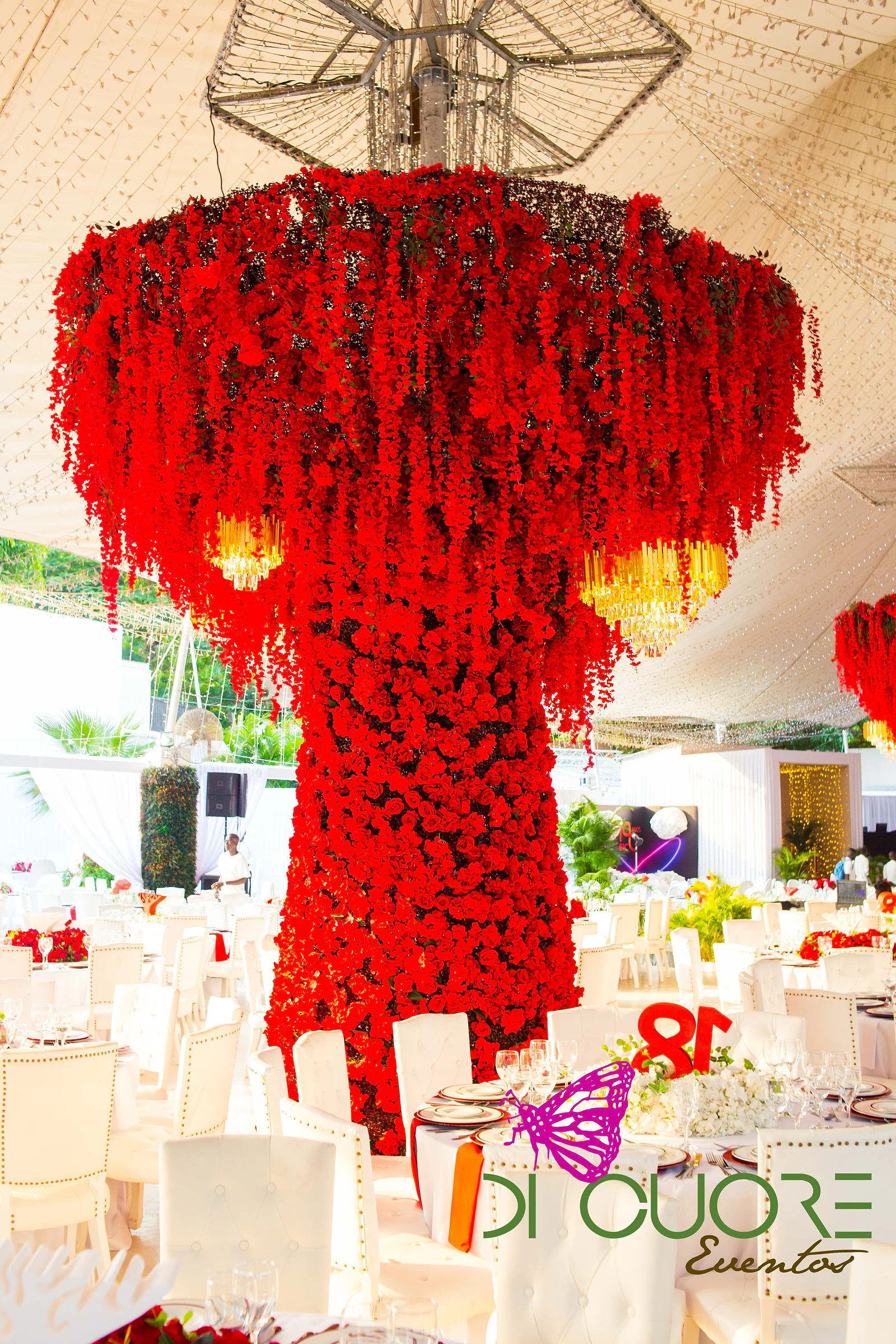 Hanging chandelier covered in cascading red flowers with golden lights, above round tables with white chairs beneath the marquee