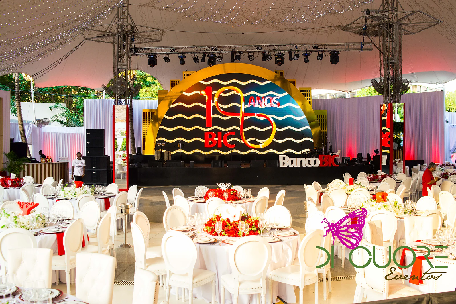 Stage with a golden arch, waves and the 18 anos BIC logo, in front of round tables with white chairs, beneath a marquee with lighting rigs