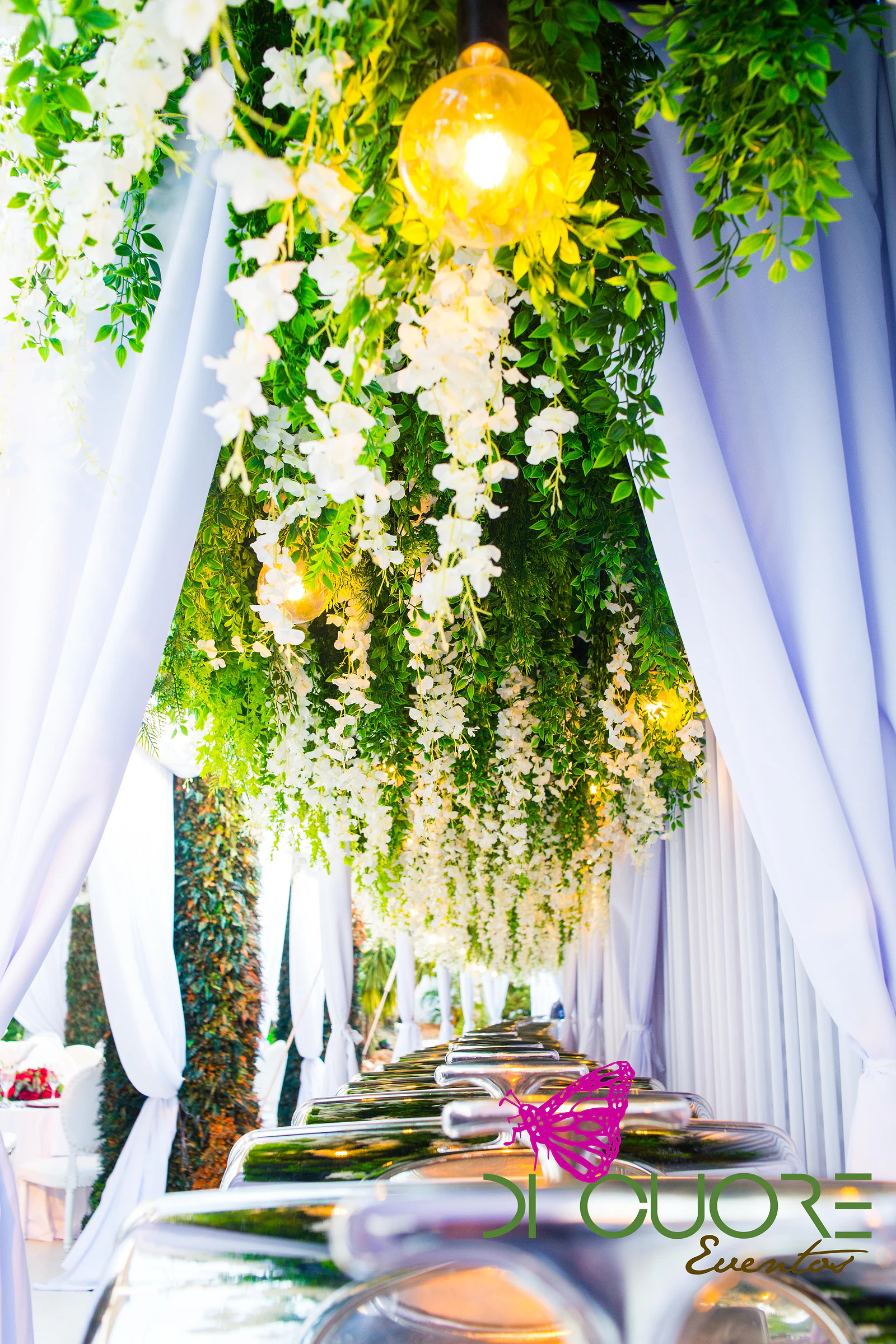 Cascade of white flowers and foliage with globe bulbs above an aisle of white drapes and buffet dishes