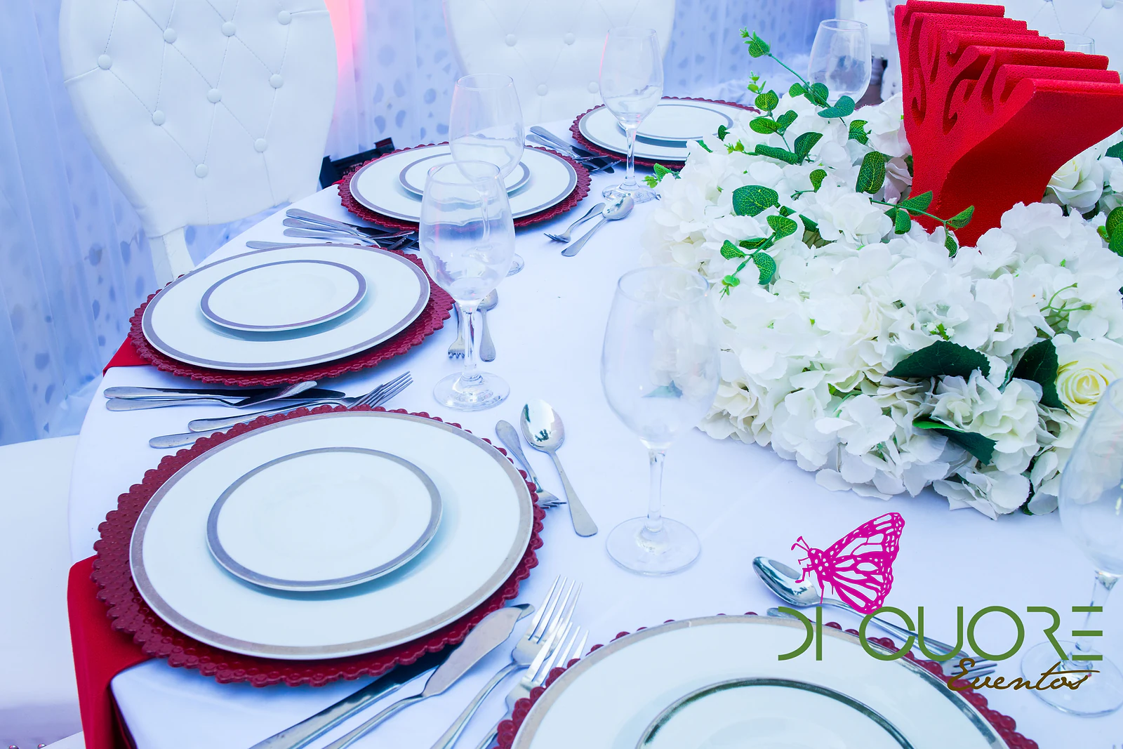 White plates with a silver rim on red chargers, with crystal glasses and a centrepiece of white hydrangeas