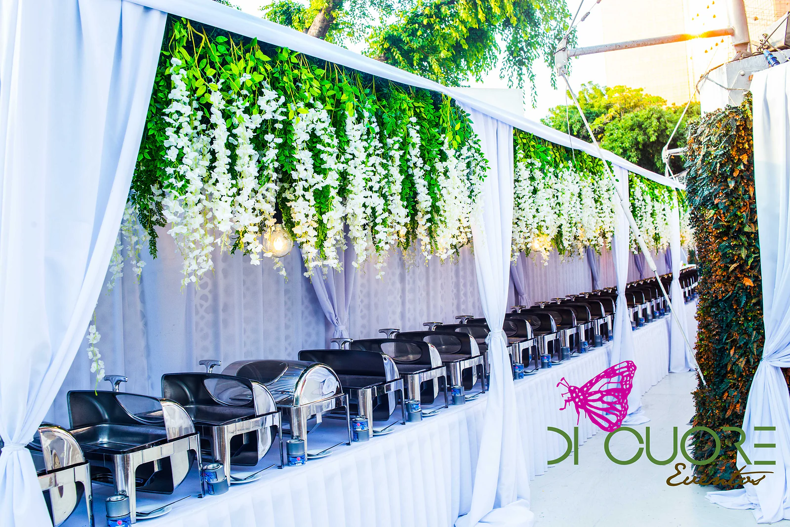 Row of buffet dishes beneath a cascade of hanging white flowers and foliage, between white drapes