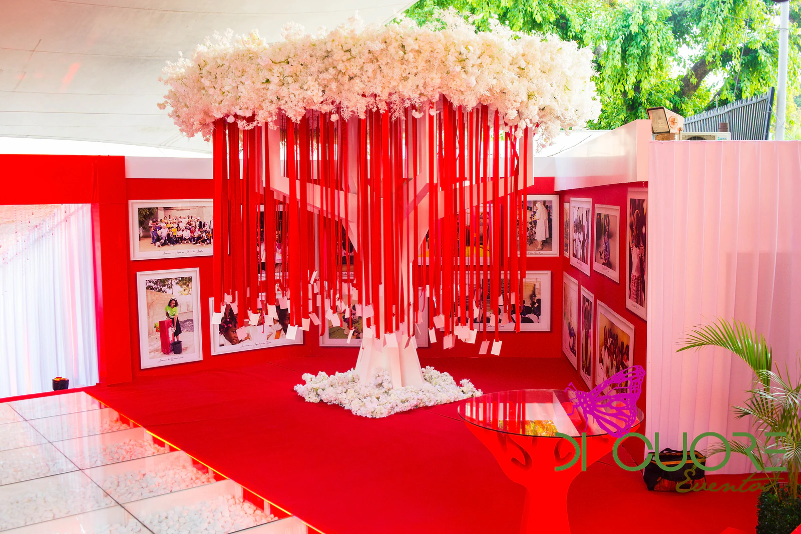 Tree of hanging red ribbons beneath a crown of white flowers, between red walls hung with framed photographs