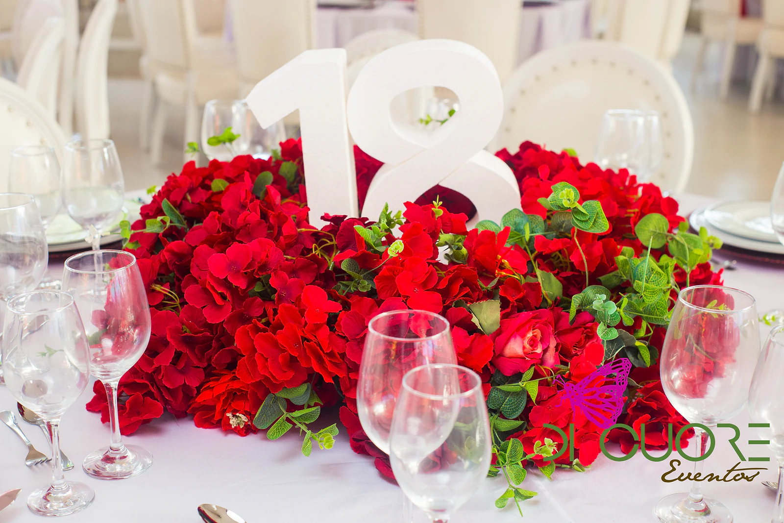 Centrepiece of red roses and hydrangeas with green leaves and a white number 18, among crystal glasses