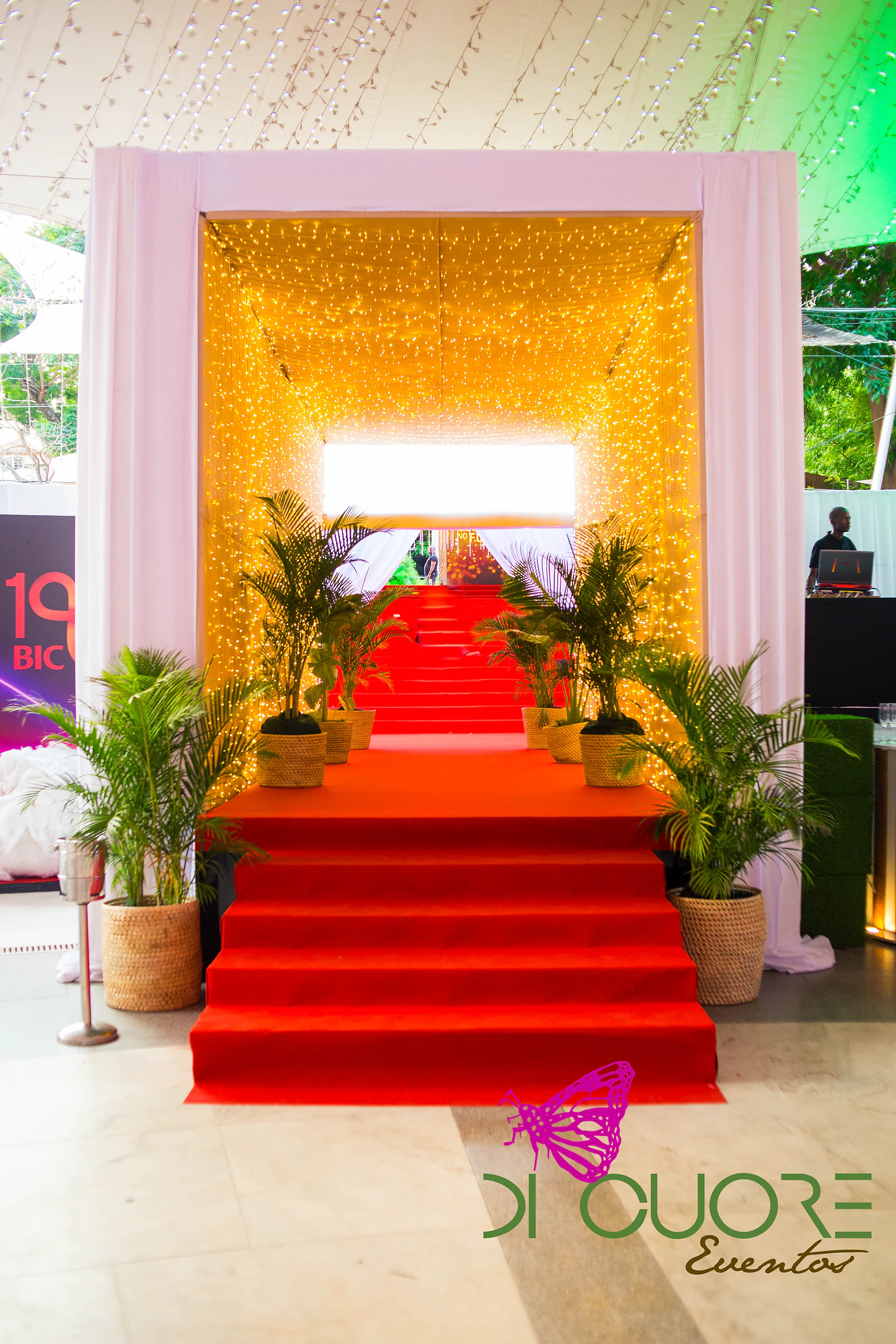 Red-carpeted steps rising to a tunnel of golden lights, flanked by palms in straw baskets and white drapes