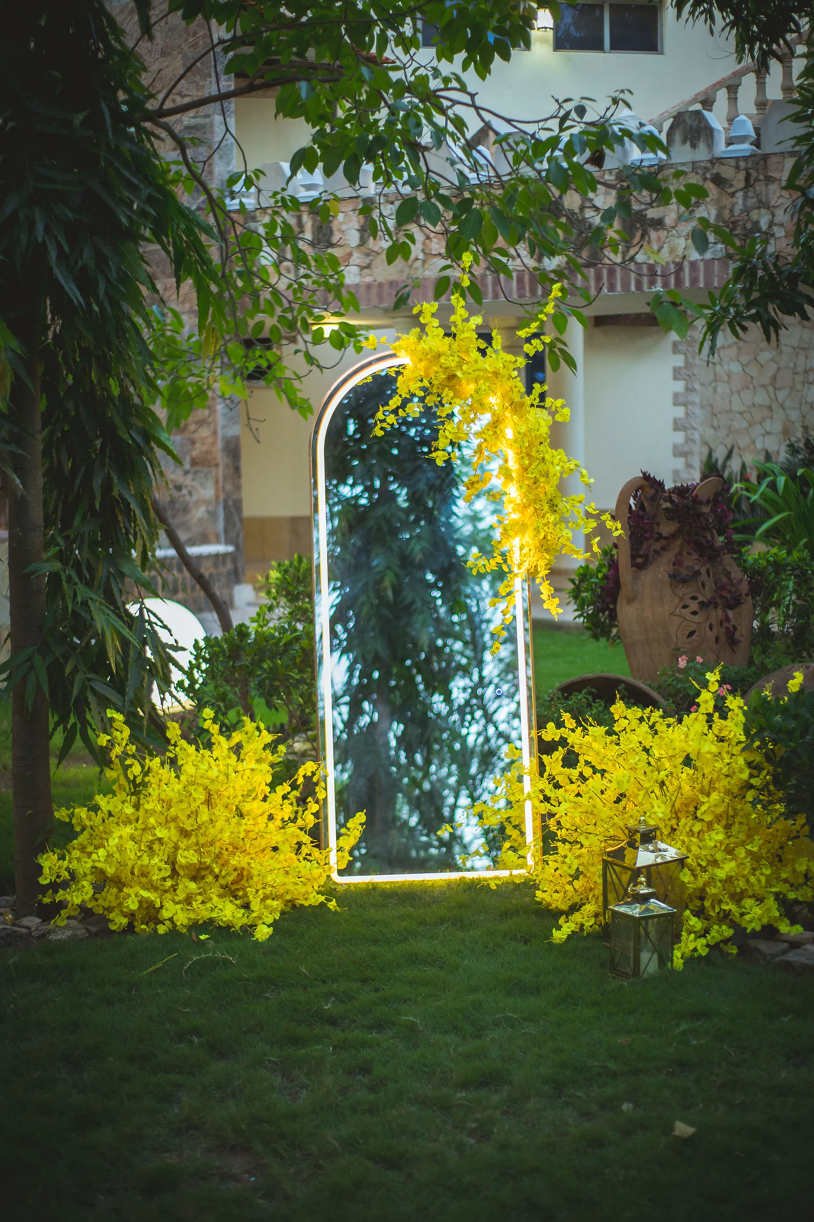 Espelho em arco com contorno de luz entre arranjos de orquídeas amarelas no relvado, com duas lanternas douradas ao lado