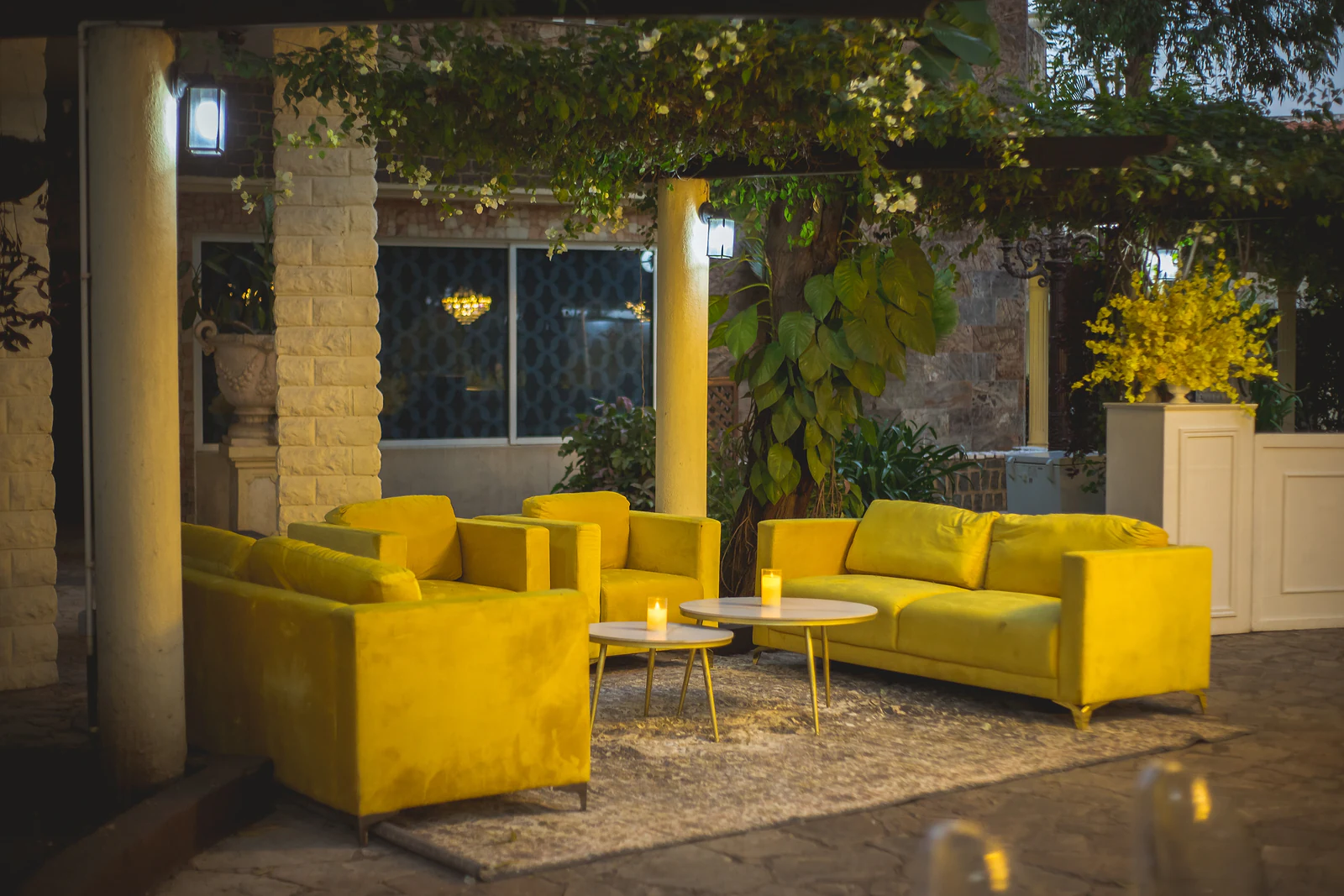 Recanto de estar com sofás e cadeirões de veludo amarelo, mesas de apoio redondas com velas e um tapete claro, sob uma pérgola