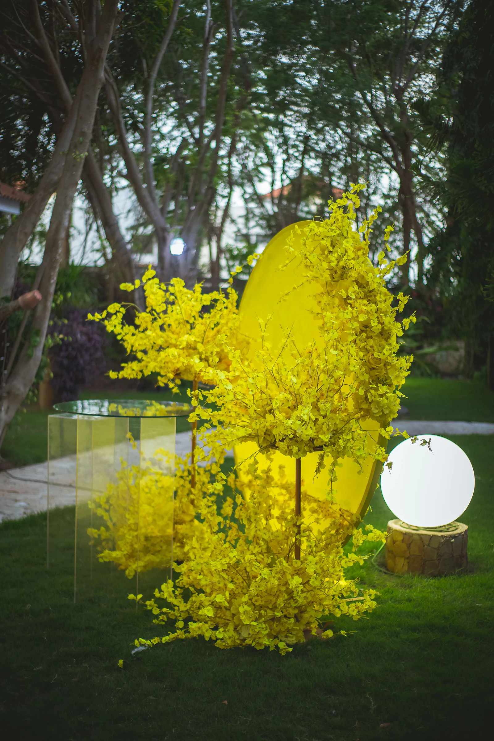 Disco amarelo coberto de orquídeas amarelas, mesa de apoio em acrílico e esfera luminosa branca sobre um pedestal de pedra, no relvado