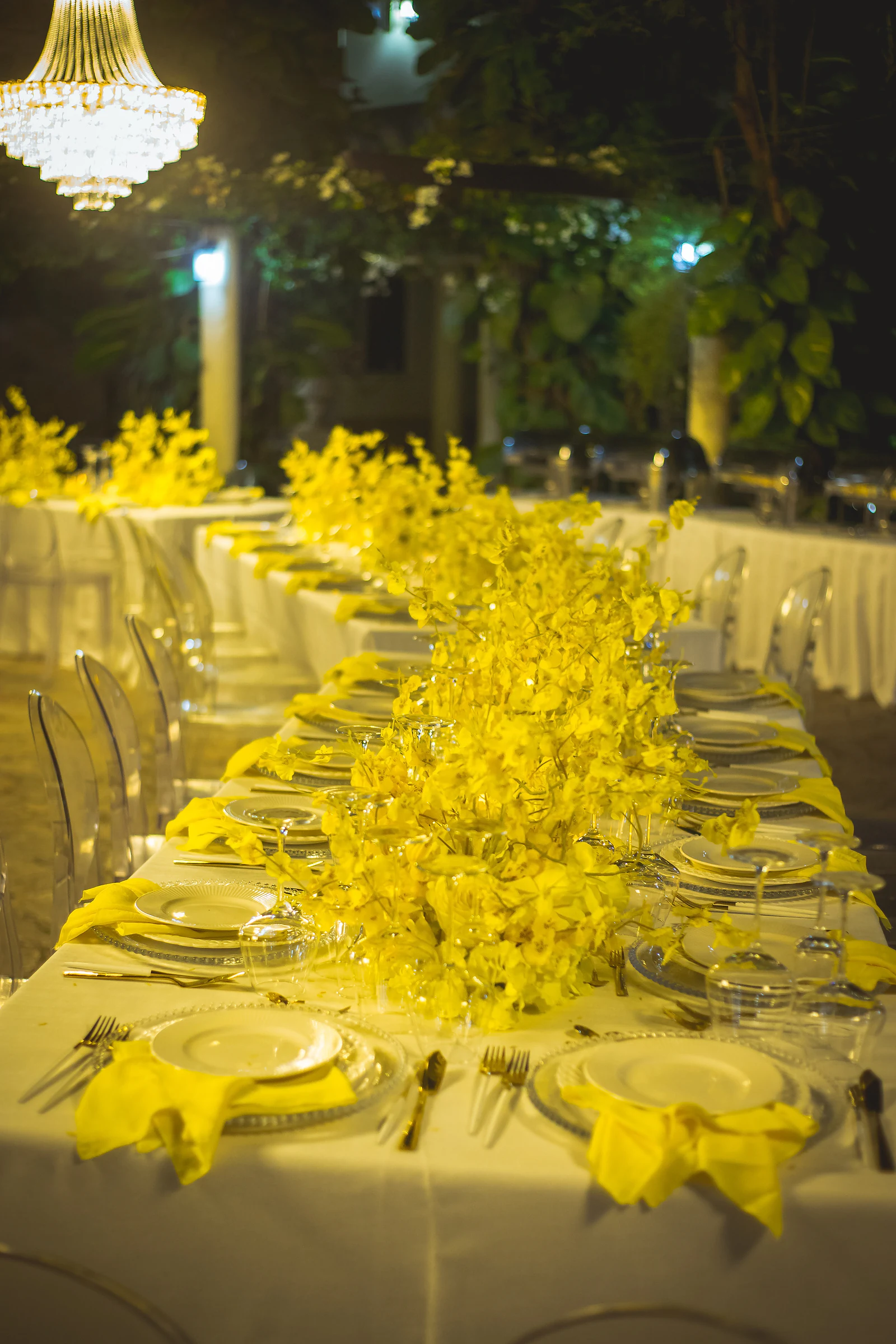 Mesa comprida com orquídeas amarelas ao centro, guardanapos amarelos sobre os pratos e um lustre de cristal no canto