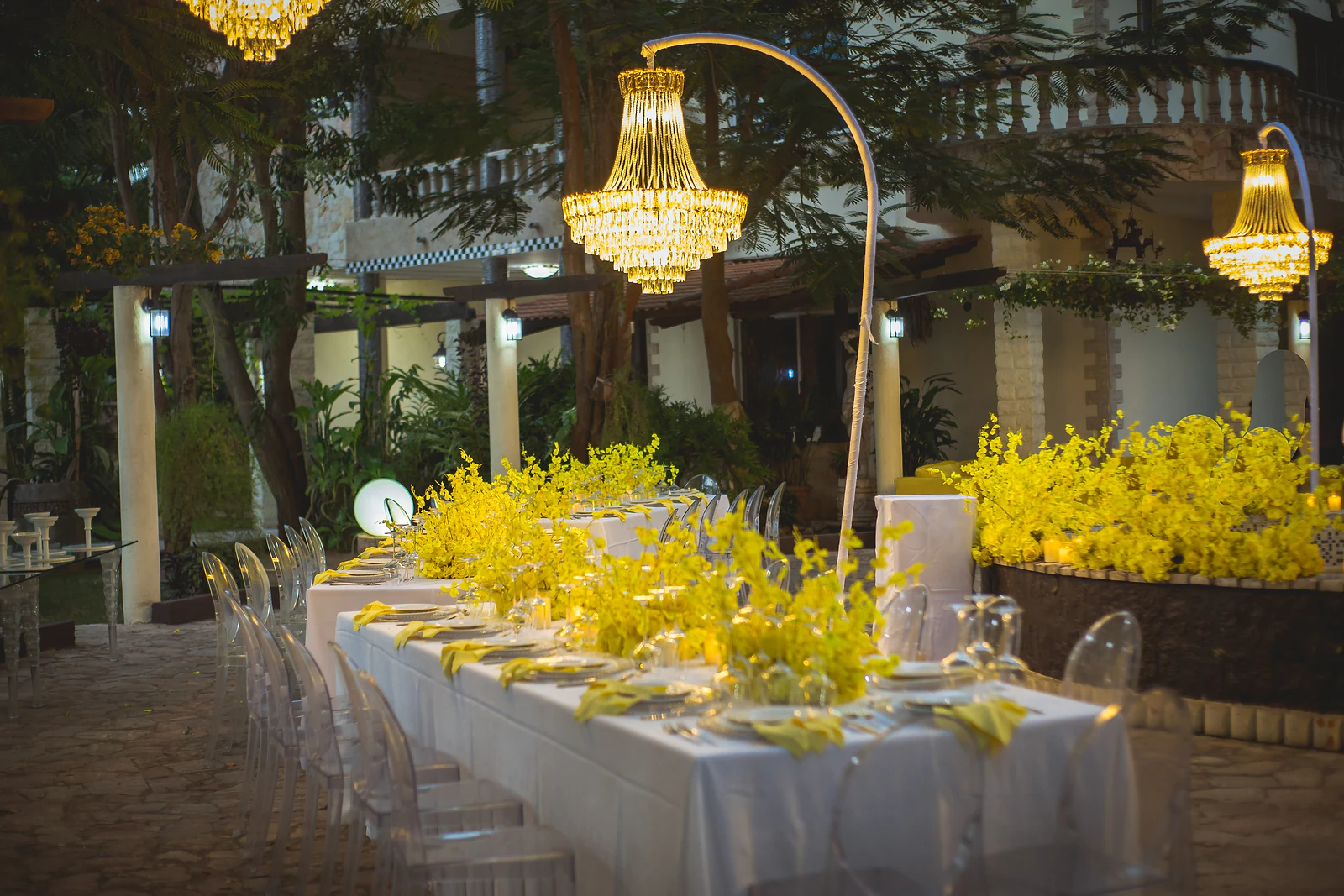 Mesa corrida de toalha branca com cadeiras transparentes e orquídeas amarelas, ao lado de um lustre de cristal dourado num jardim ao anoitecer