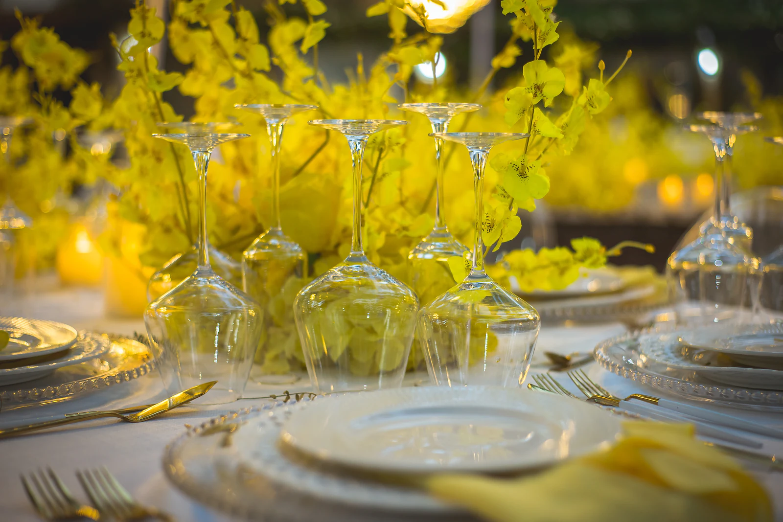 Taças de cristal, pratos de rebordo de contas e talheres dourados diante de um centro de mesa de orquídeas amarelas e velas
