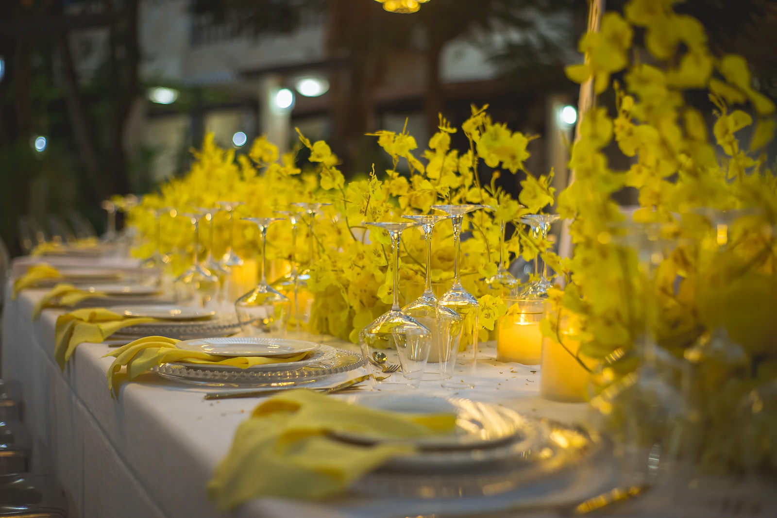Fila de pratos com guardanapos amarelos, velas e orquídeas amarelas ao longo da mesa comprida