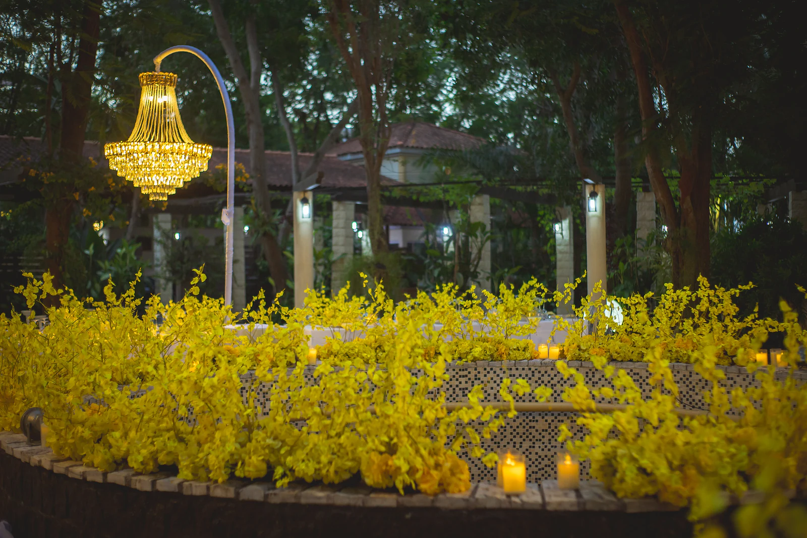 Floreira circular coberta de orquídeas amarelas e velas, com um lustre de cristal em haste branca e o jardim ao fundo