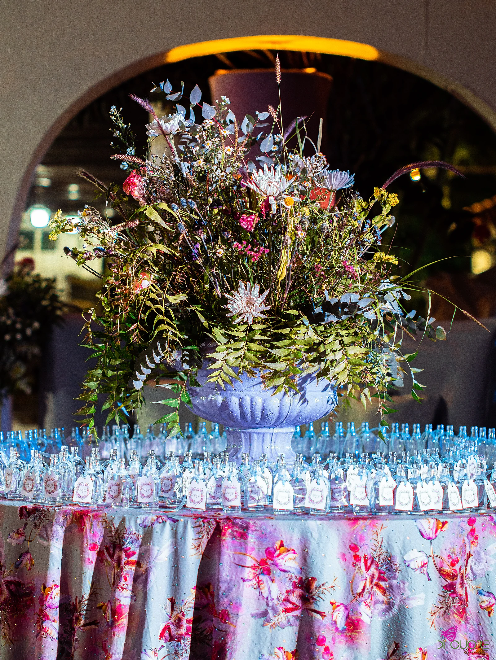 Grande arranjo de flores silvestres numa urna lilás, rodeado de pequenos frascos de vidro com etiquetas, sobre toalha estampada a flores, diante de um arco iluminado