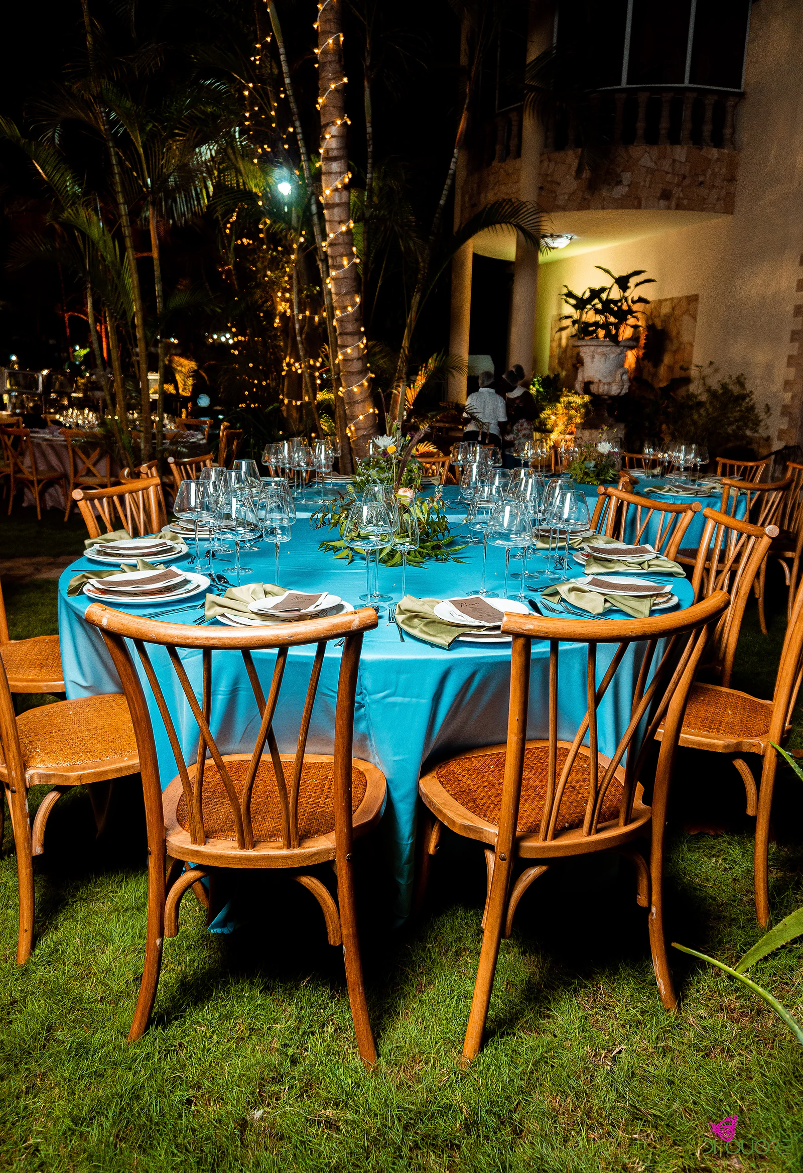 Round table with a turquoise cloth set for dinner, with wooden chairs on grass and a palm trunk wound with fairy lights beyond