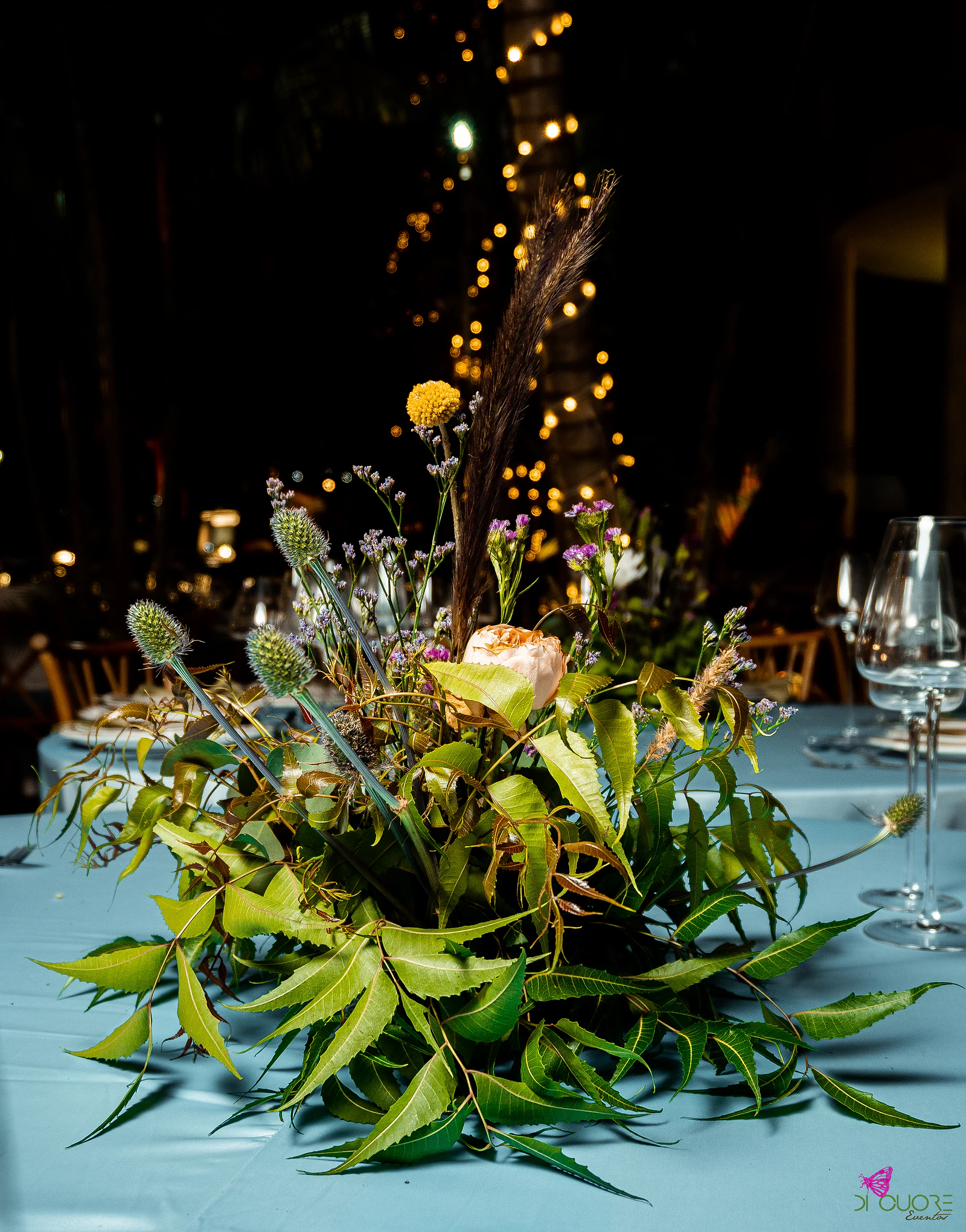 Low foliage centrepiece with blue thistles, a pale rose, a round yellow flower and a brown plume on a pale blue cloth, with lights beyond