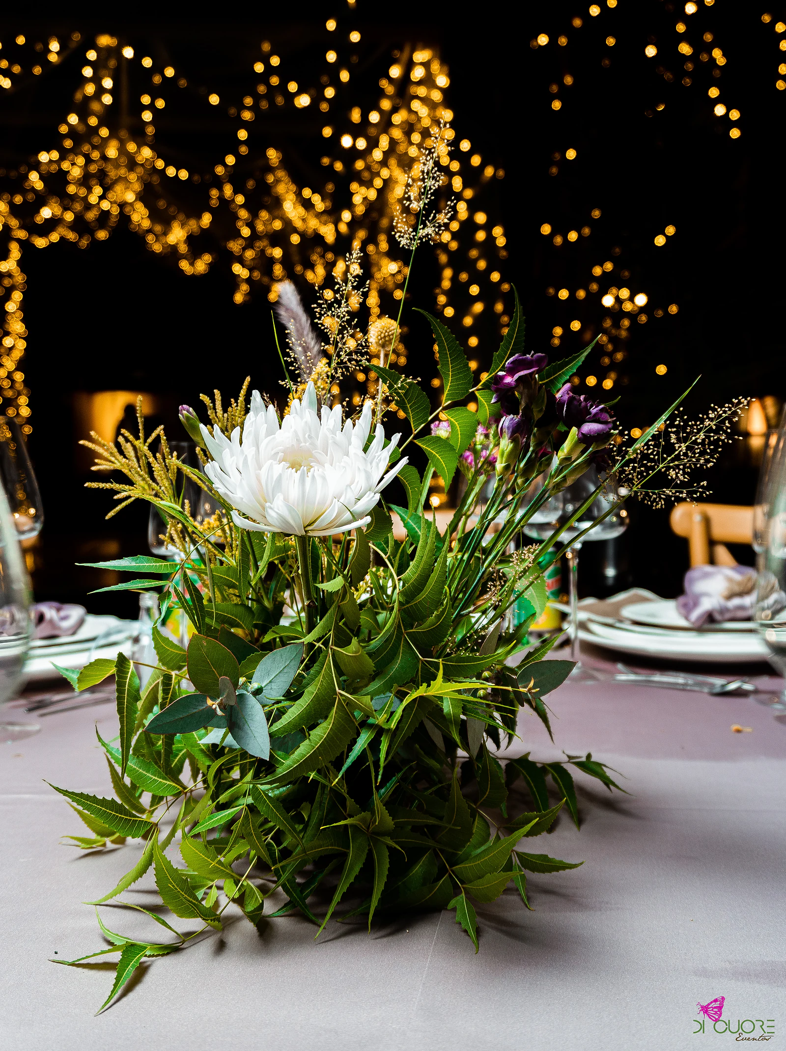 Foliage centrepiece with a white bloom on a pale cloth, with soft-focus lights behind