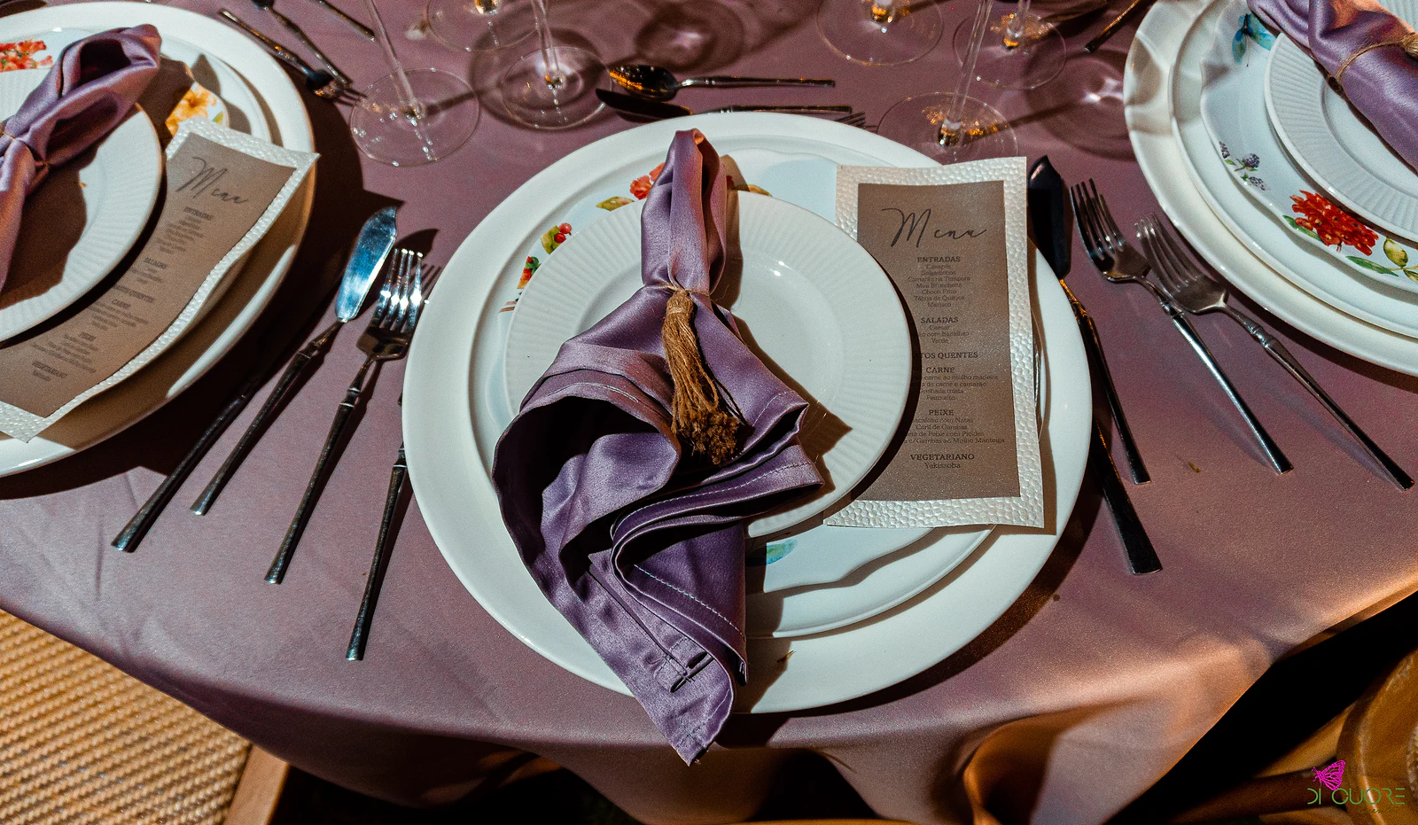 White plate with a lilac napkin tied with a tasselled cord and a kraft-paper menu, on a mauve cloth, beside plates painted with flowers