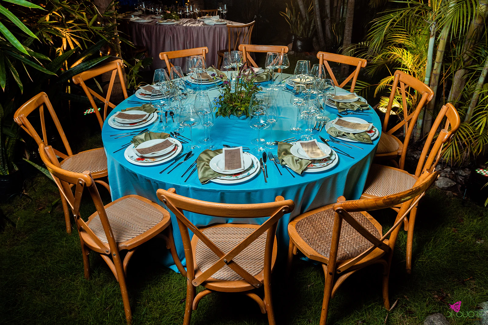 Round table with a turquoise cloth, cane-seated wooden chairs and a foliage centrepiece, on grass