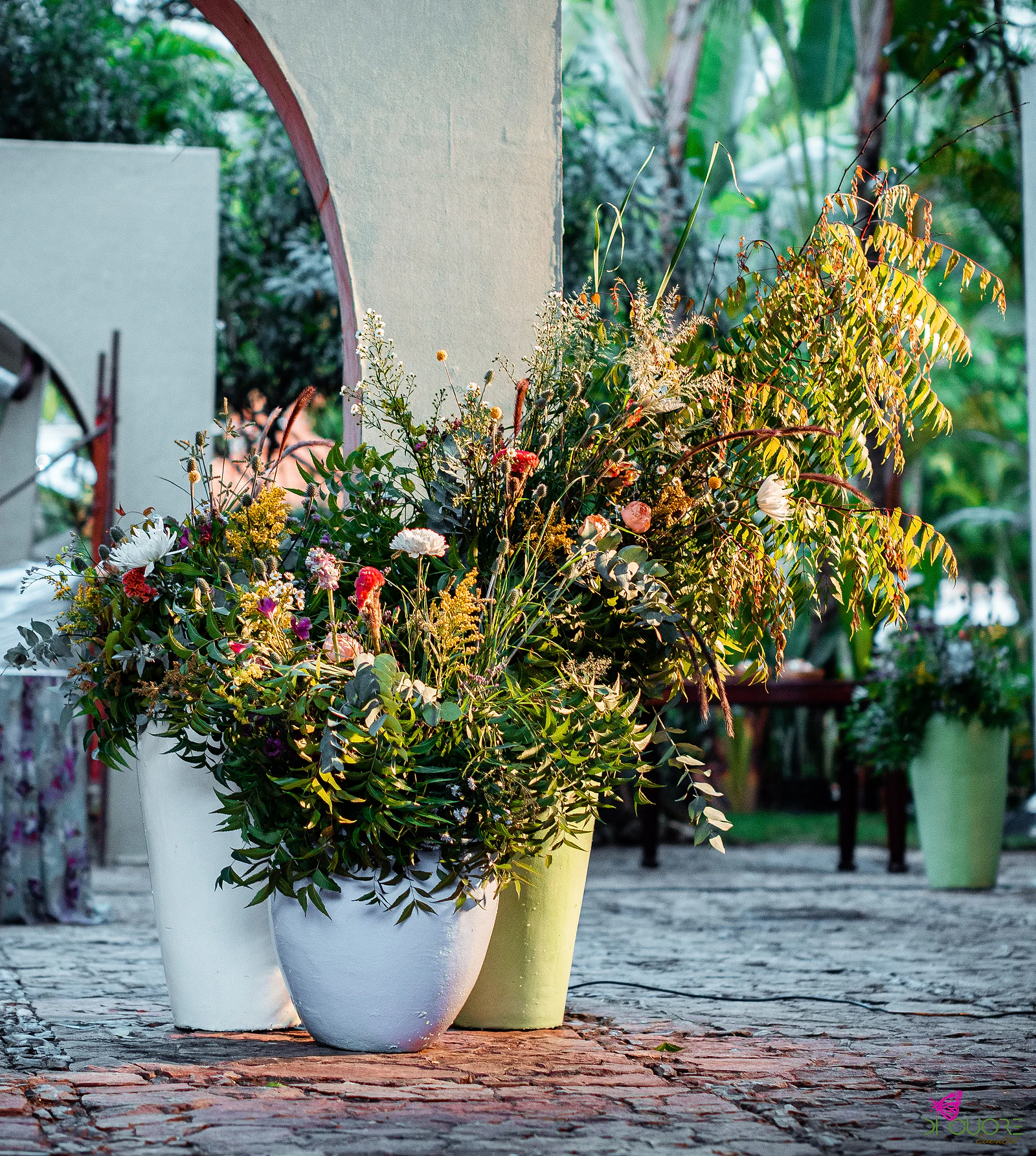 Three vases, two white and one lime green, overflowing with wildflowers and foliage beside a white arch, on a cobbled pavement