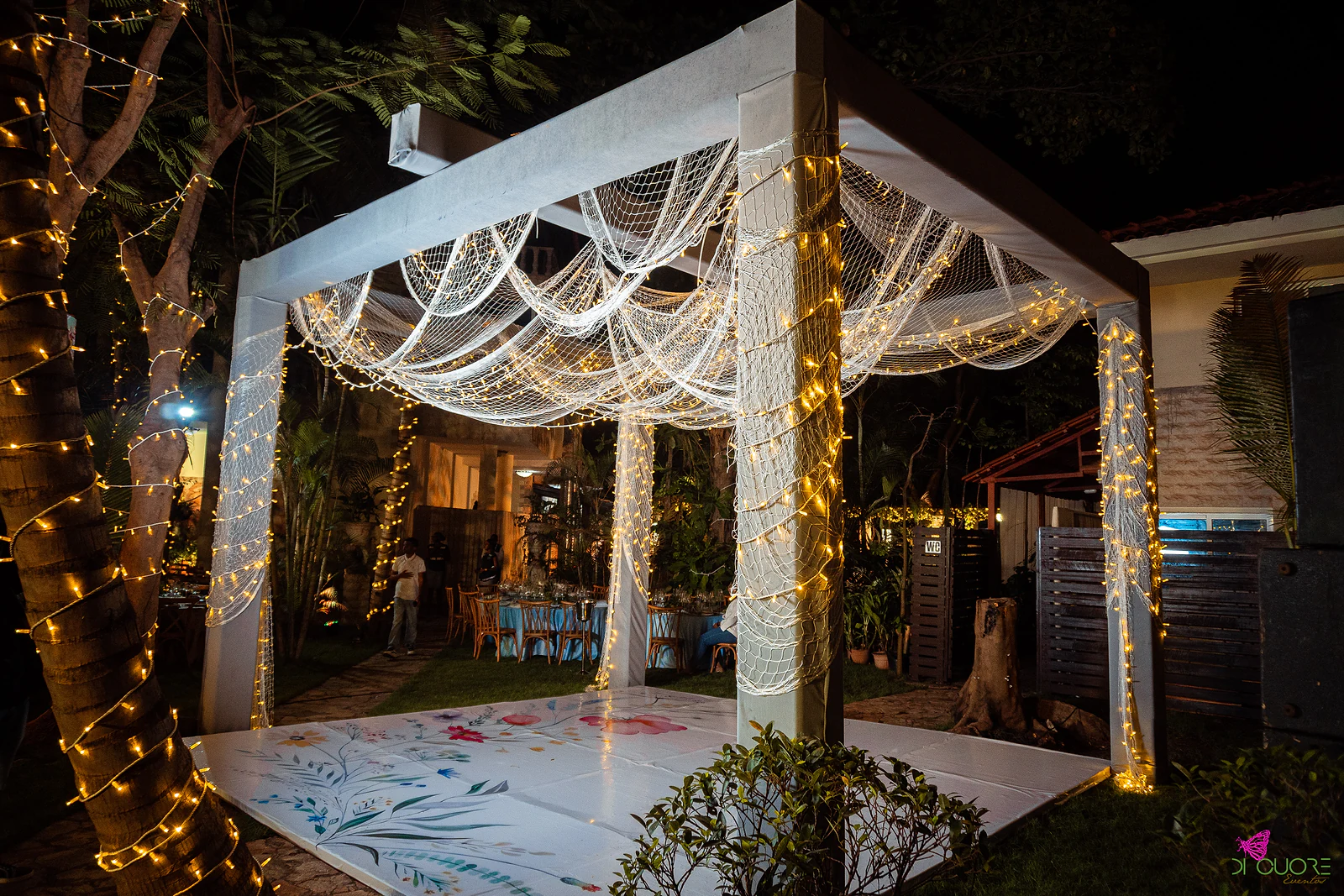 White gazebo draped in fairy lights over a white dance floor with a floral design, among lit trees