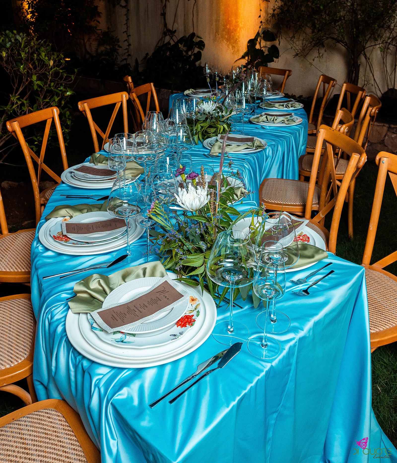 Long table with a turquoise satin cloth, white plates painted with flowers, olive-green napkins, kraft menus and a foliage centrepiece with white blooms, among wooden chairs