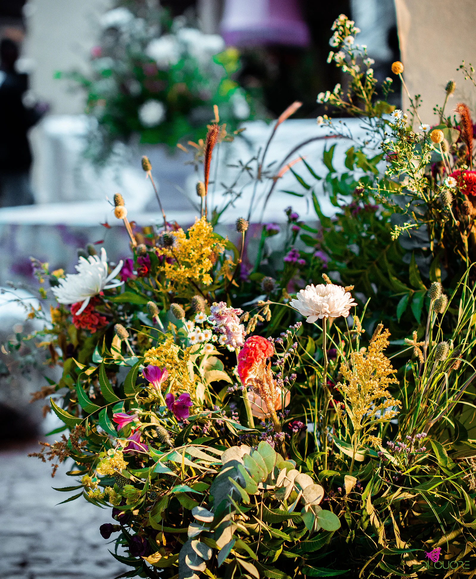 Close-up of yellow, red, pink and white wildflowers with eucalyptus leaves, before a soft-focus table