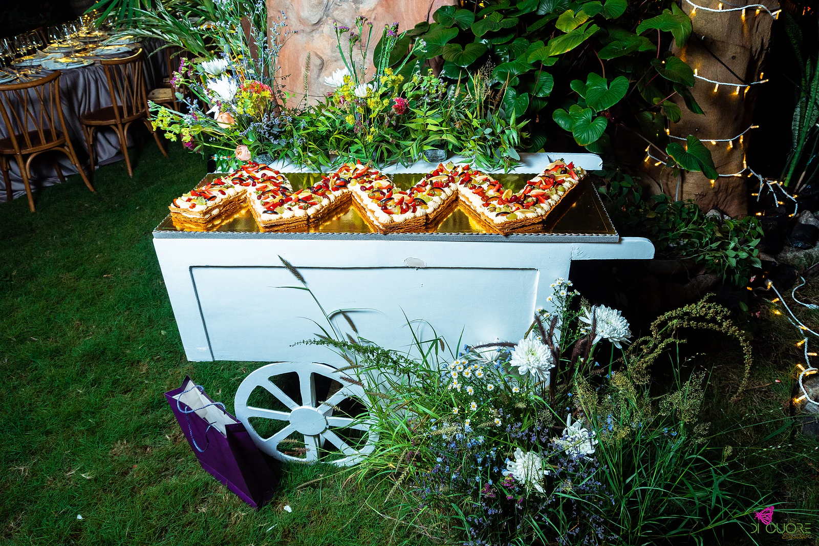 White cart with slices of layered pastry with cream and red fruit on a gold tray, among white wildflowers and foliage, on grass and fairy lights