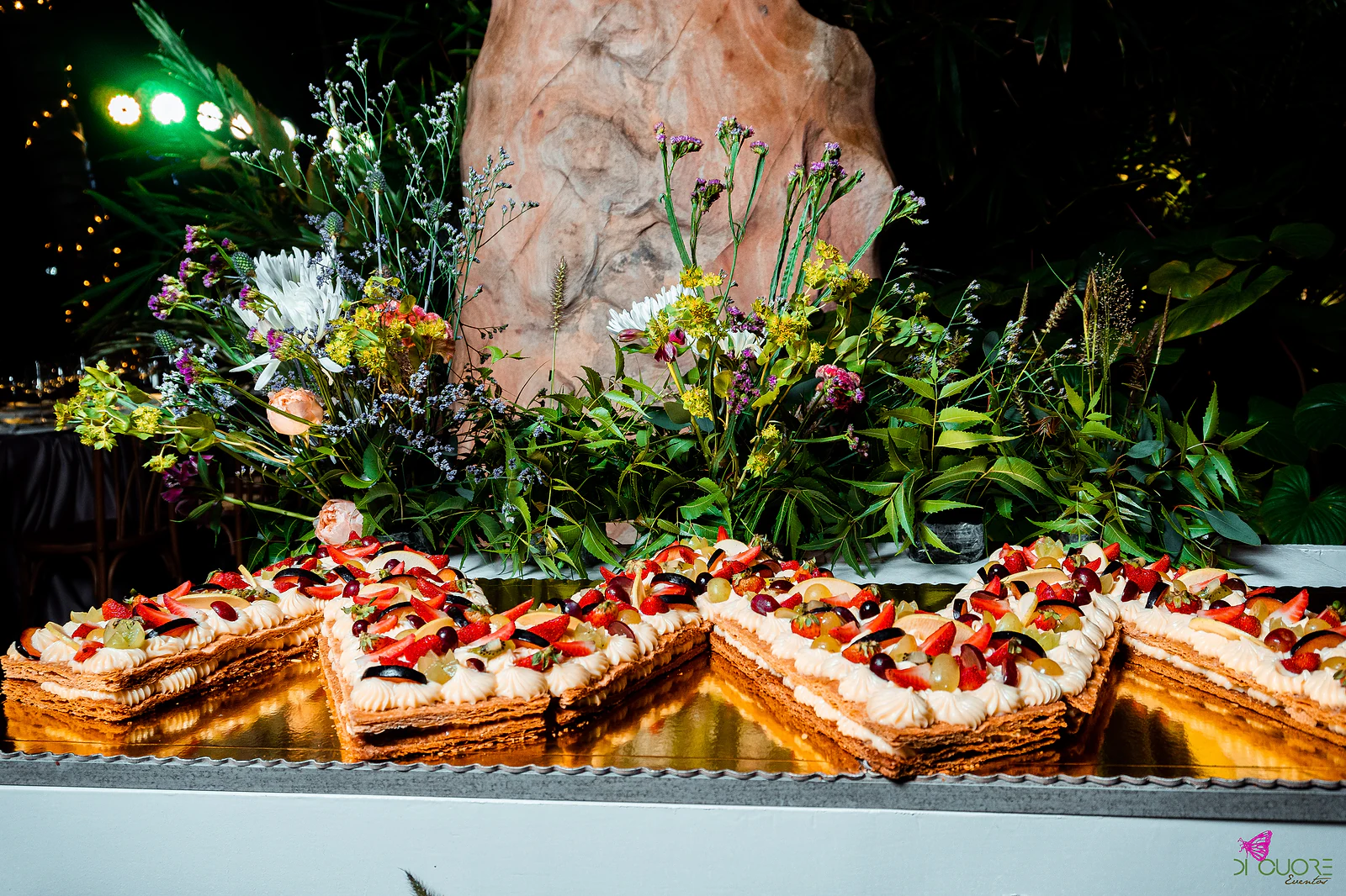 Slices of layered pastry with cream and red fruit on a gold tray, surrounded by wildflowers and foliage