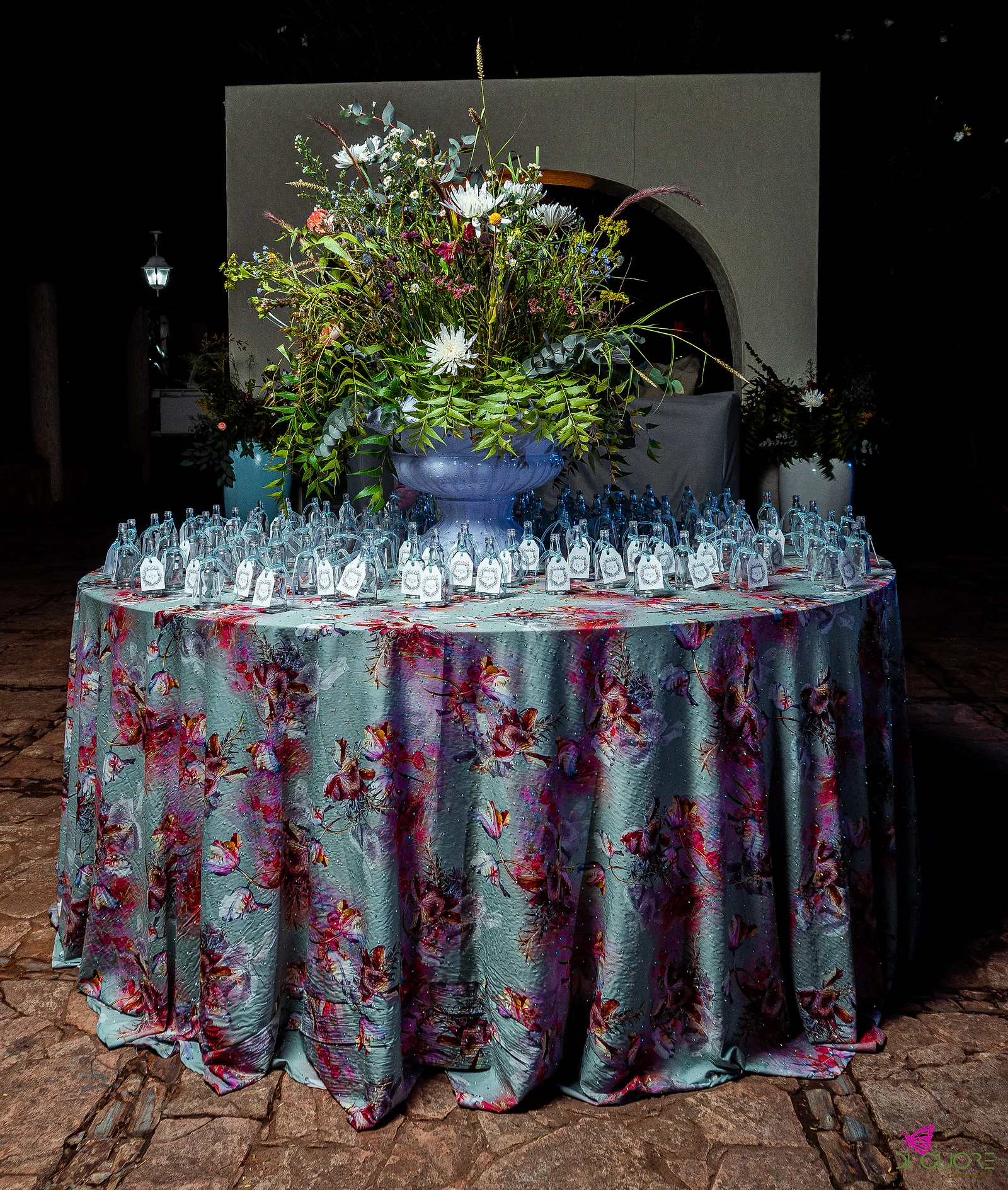 Round table with a floral-print cloth and small labelled glass bottles beneath a large arrangement of wildflowers in a lilac urn, before an arch