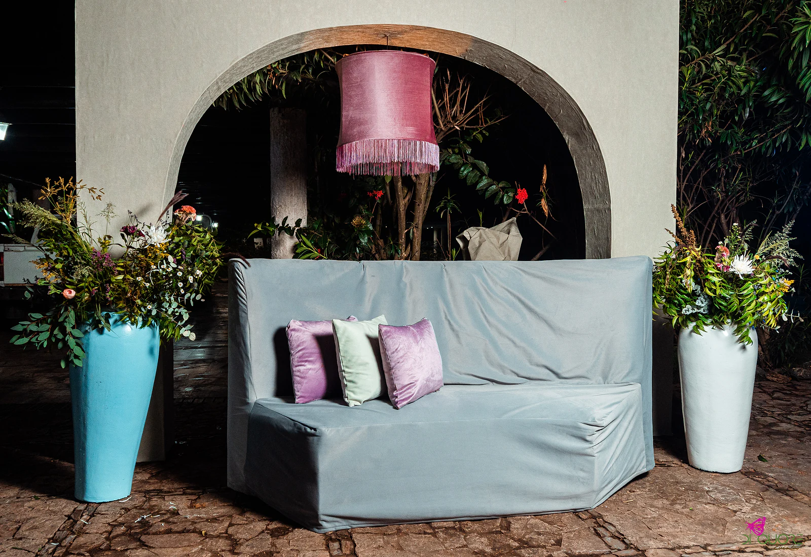 Grey sofa with lilac and green cushions between tall vases of flowers, beneath an arch with a hanging pink lampshade