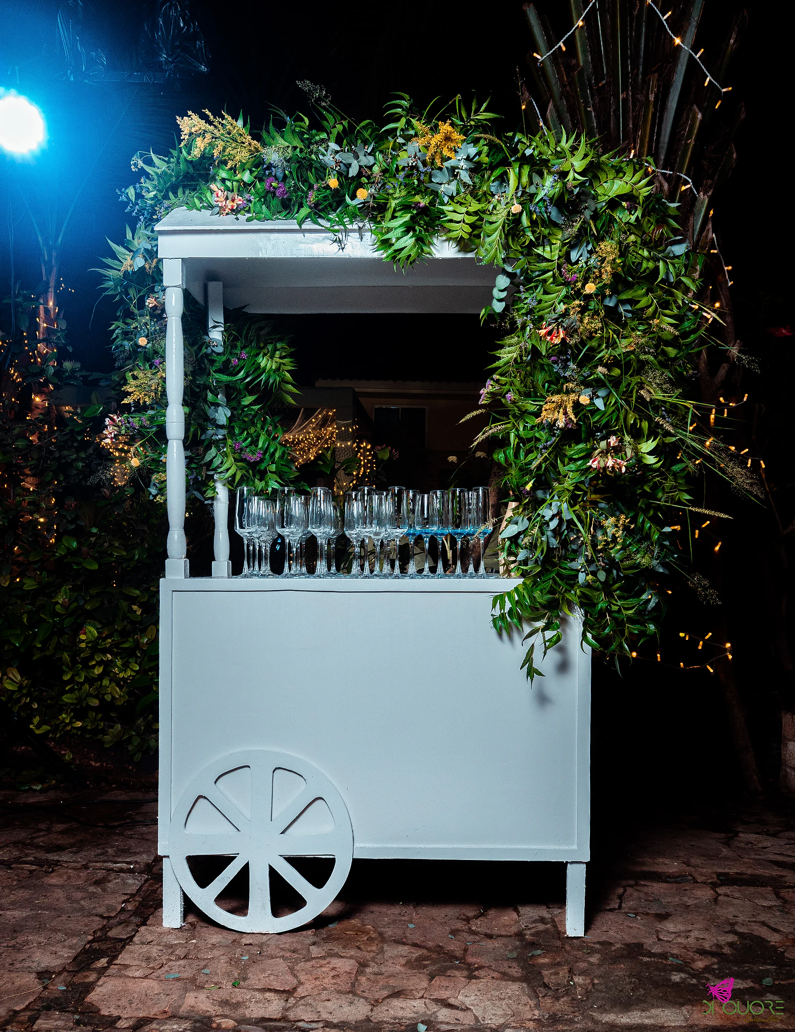 White cart with a garland of foliage and wildflowers and a row of glasses on the counter