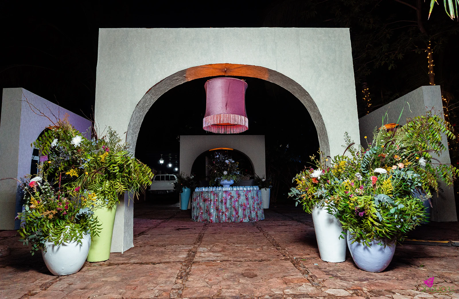White arched entrance with a hanging pink fringed lampshade between large vases of wildflowers, and a floral-print table beyond, at night