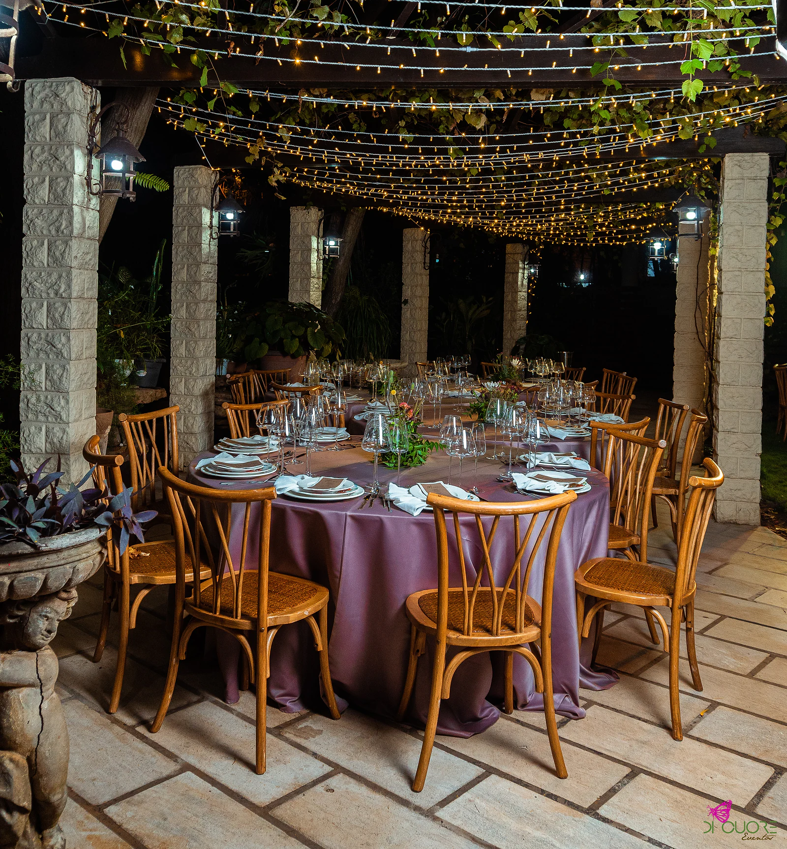 Round table with a lilac cloth set for dinner beneath a stone-pillared pergola hung with fairy lights, with wooden chairs and a carved stone urn in the foreground