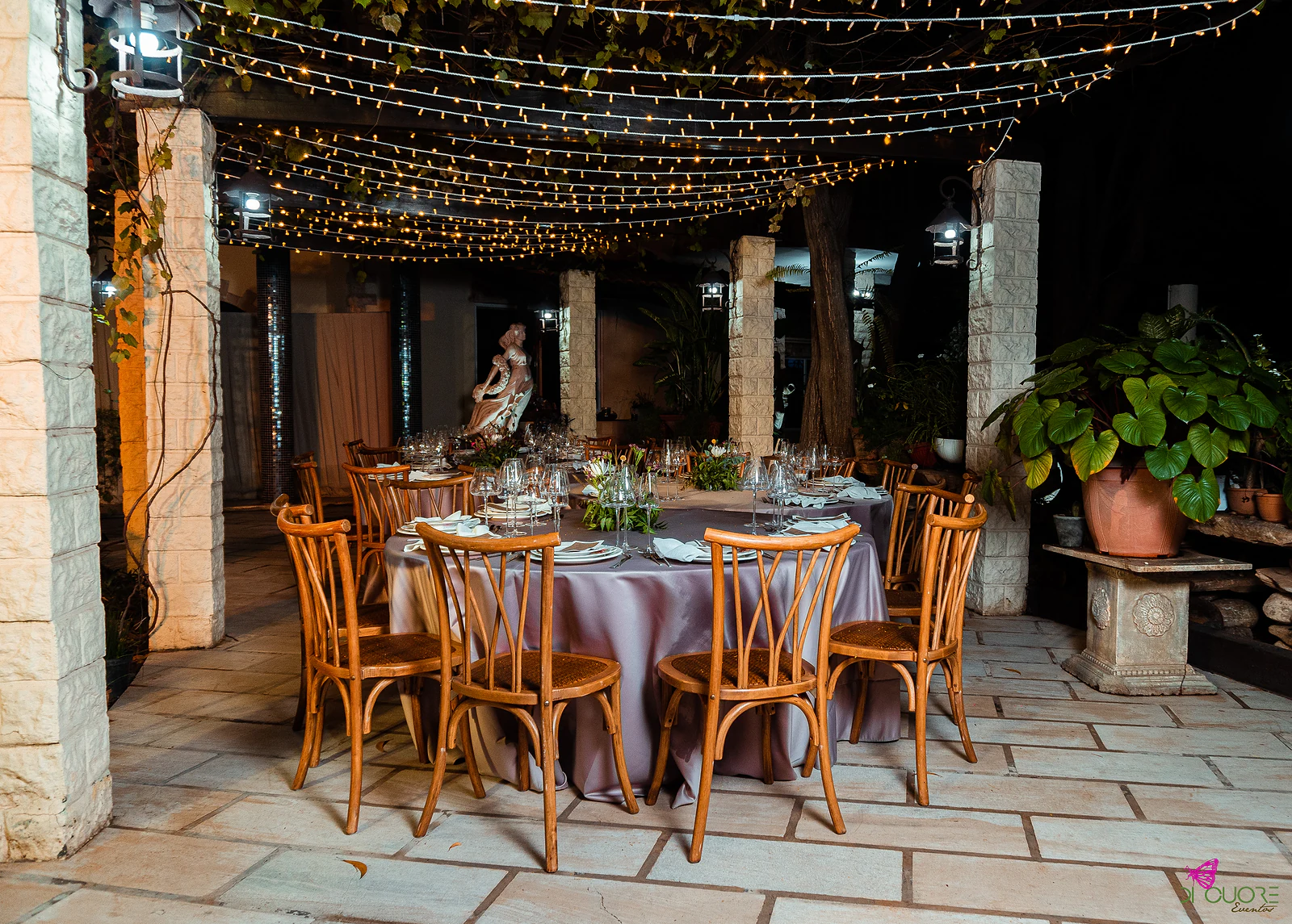 Round table with a lilac cloth beneath a pergola hung with fairy lights, with wooden chairs