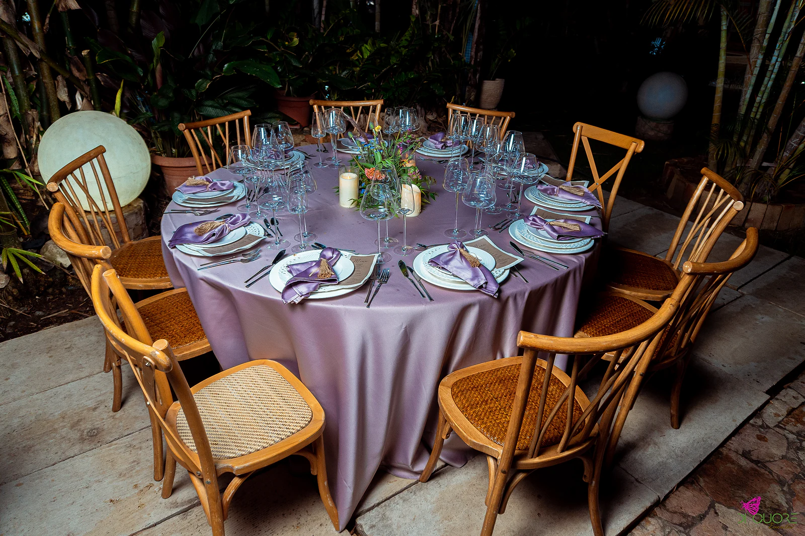 Round table with a lilac cloth, purple napkins, white candles and a foliage centrepiece, on a plank floor, with wooden chairs and a glowing globe beyond