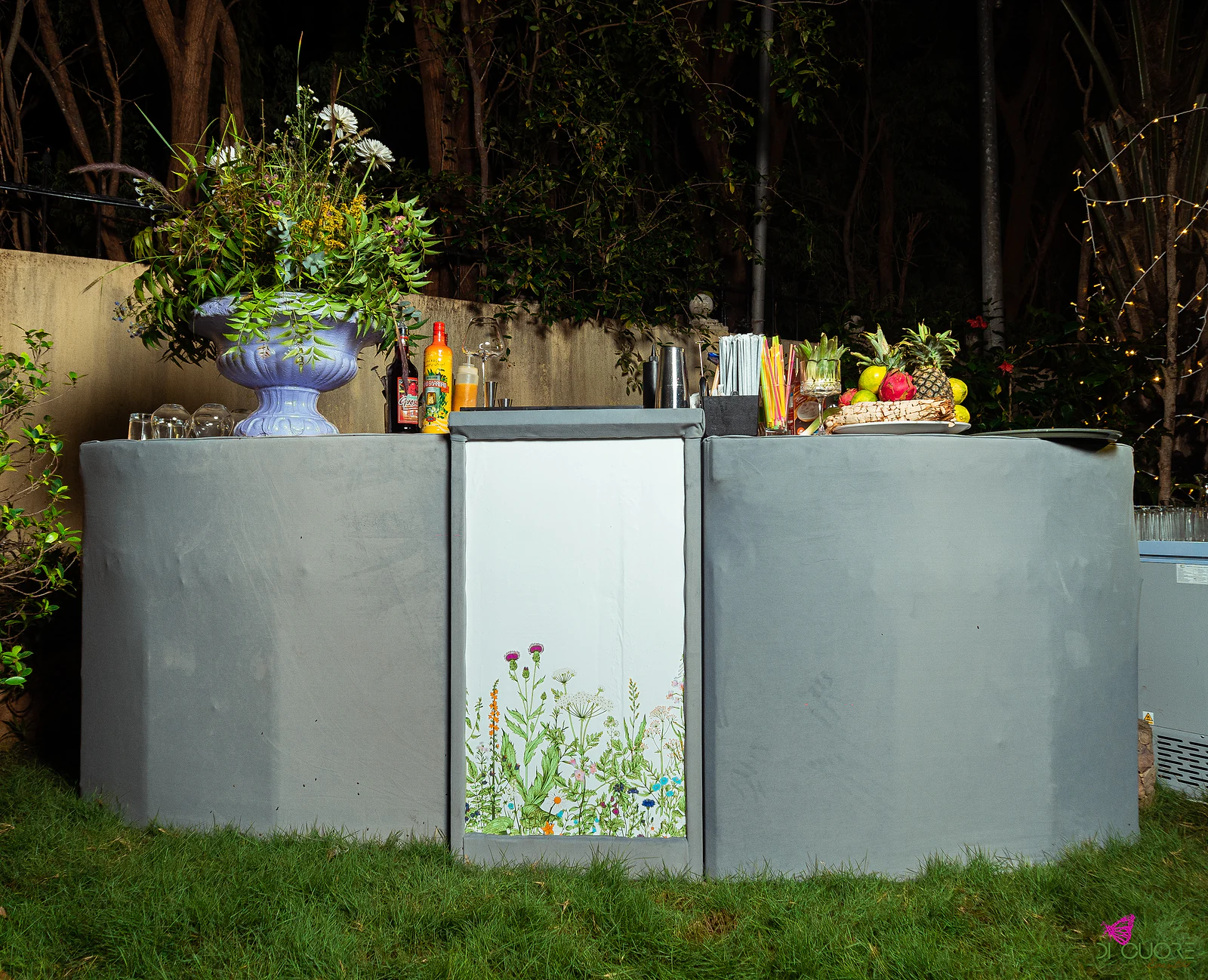 Bar counter clad in grey, with a white panel of wildflowers at the centre, a lilac urn of flowers on top, bottles, glasses and fruit, on grass