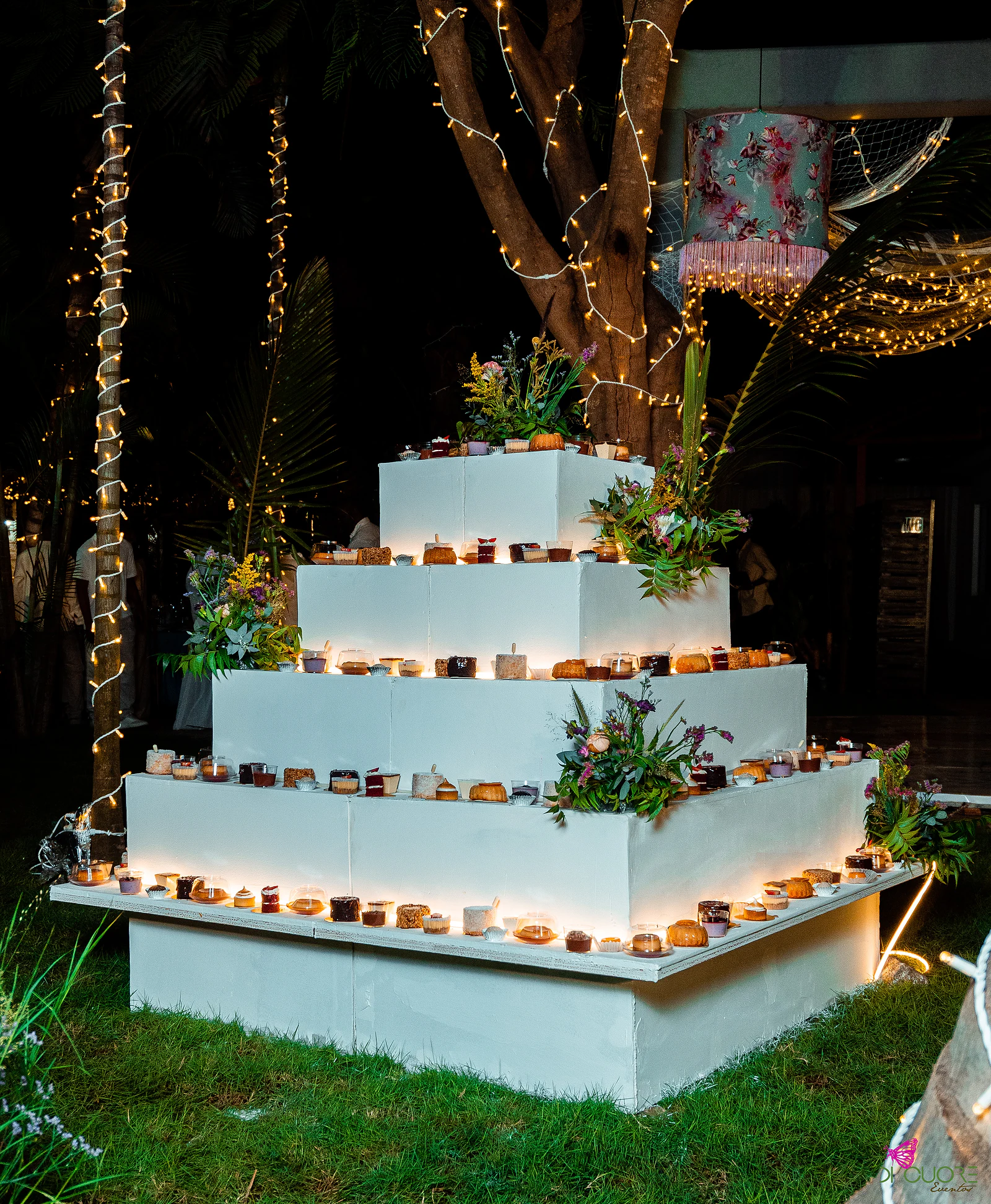 Four-tier white pyramid of small desserts and wildflowers, lit by strings of lights, beside a tree and a floral lampshade