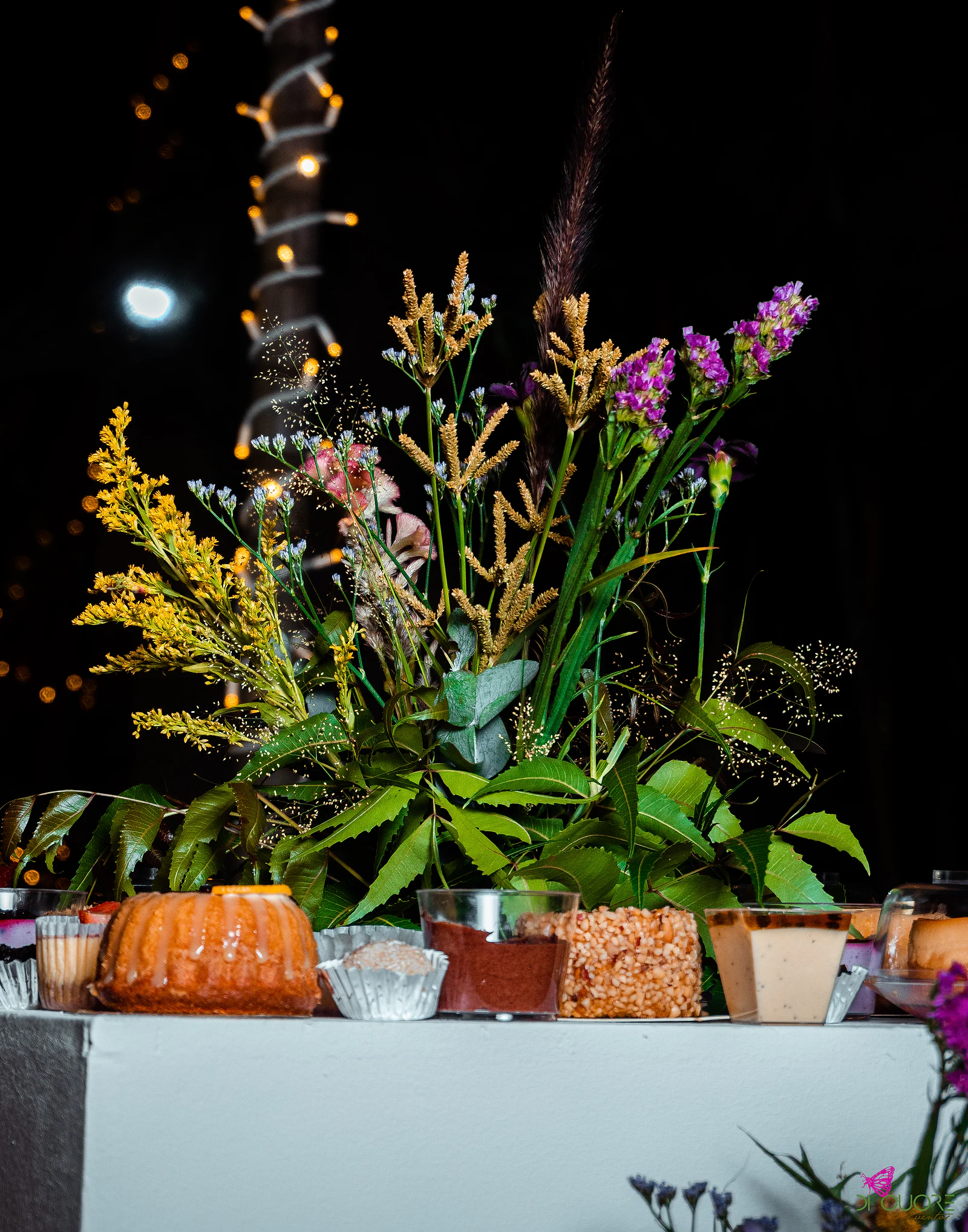 White dessert table with a cake and small cups beneath a tall arrangement of yellow, lilac and pink wildflowers