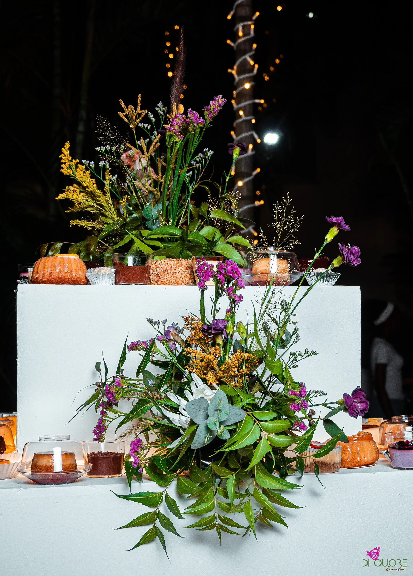 White two-level dessert table with arrangements of lilac, yellow and pink wildflowers, foliage and a succulent, among small cakes and cups