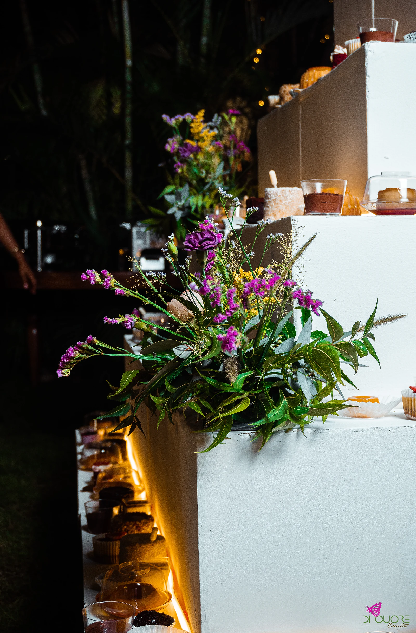 Side view of a white stepped dessert structure, with lilac wildflowers and foliage in the foreground and a row of sweets lit by a warm glow