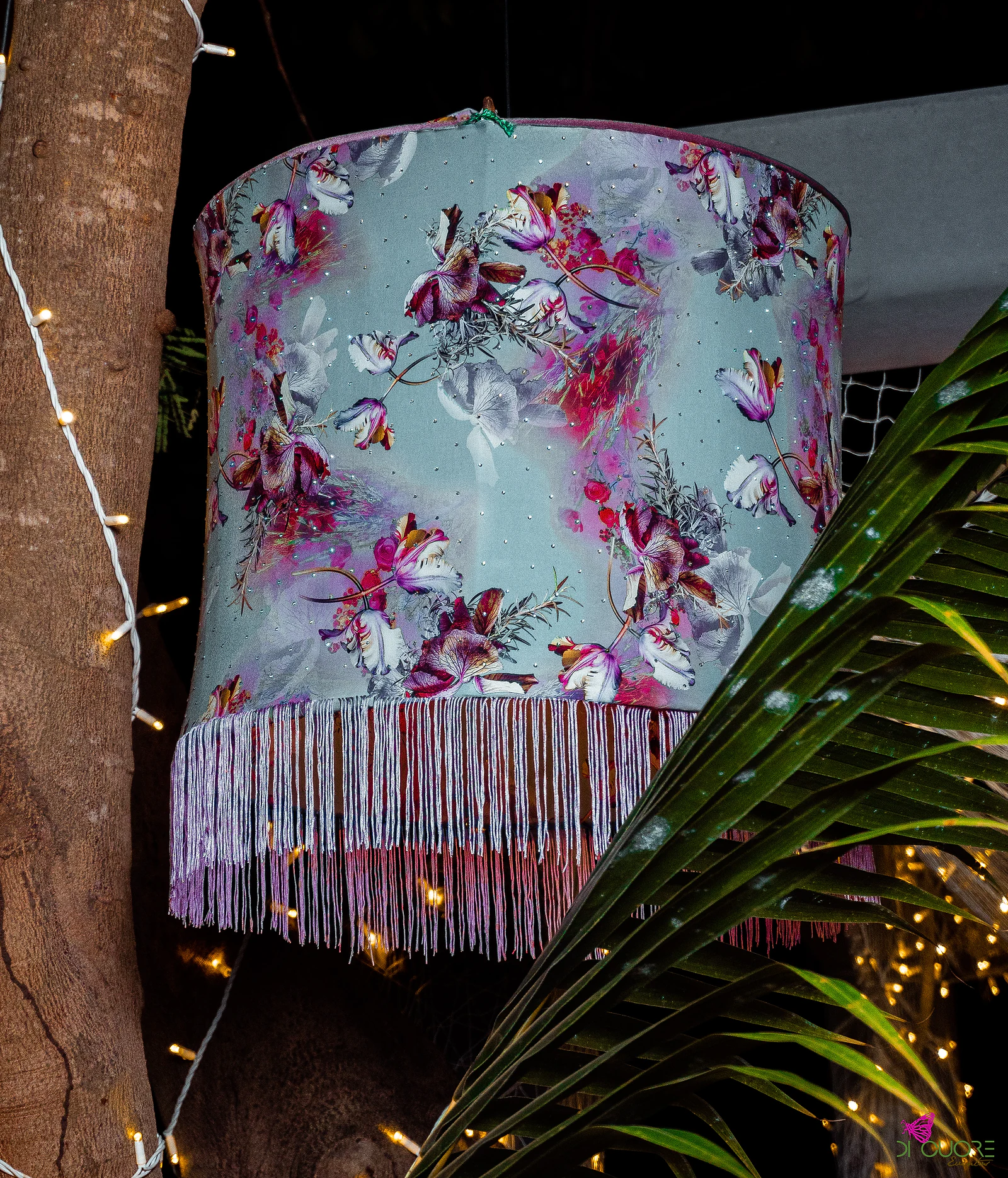 Lampshade printed with pink flowers and edged with lilac fringe, hanging among fairy lights and a palm