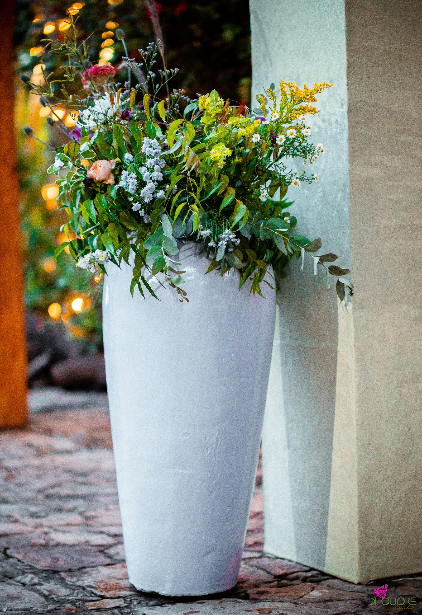 Tall white vase of yellow, white and pink wildflowers beside a column
