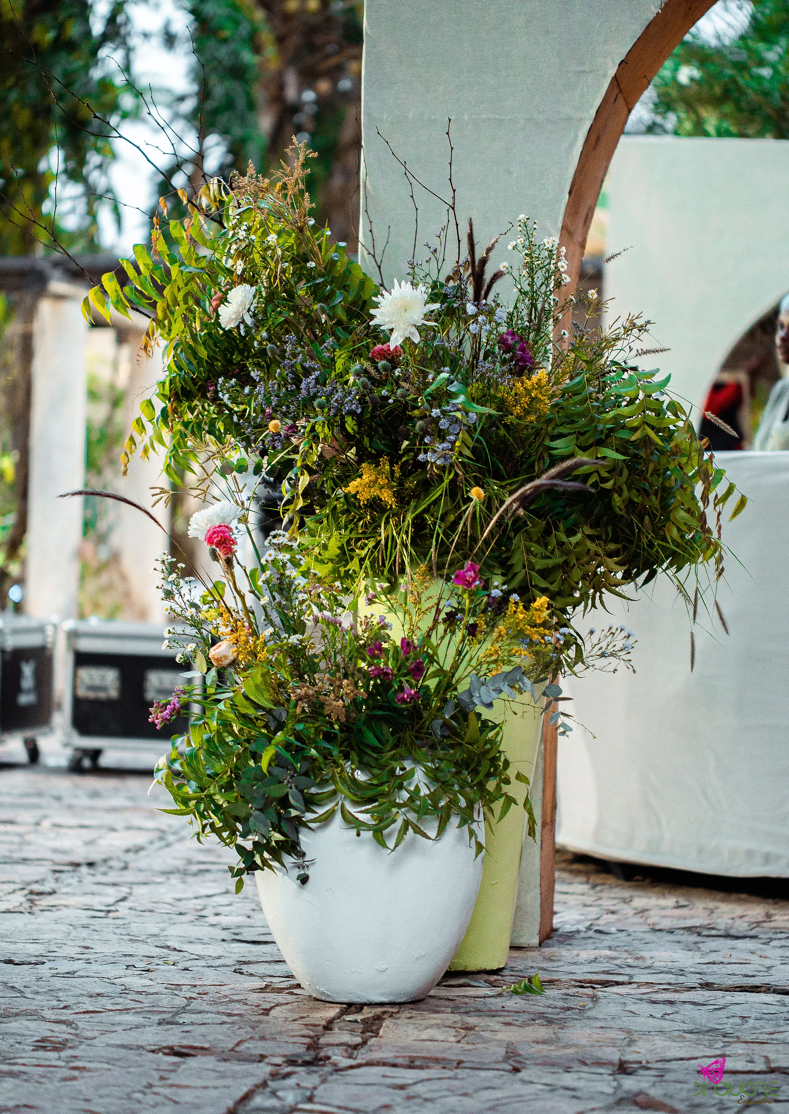 Two vases, one white and one lime green, holding a tall arrangement of foliage and white, pink and yellow wildflowers beside a white arch