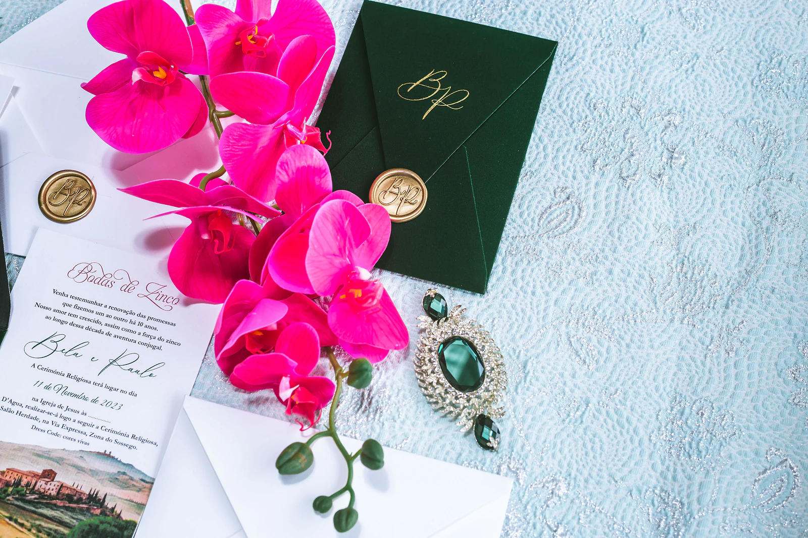 A white invitation card beside a dark green envelope with a gold monogram and seal, pink orchids and a brooch of green stones, on pale blue embroidered fabric