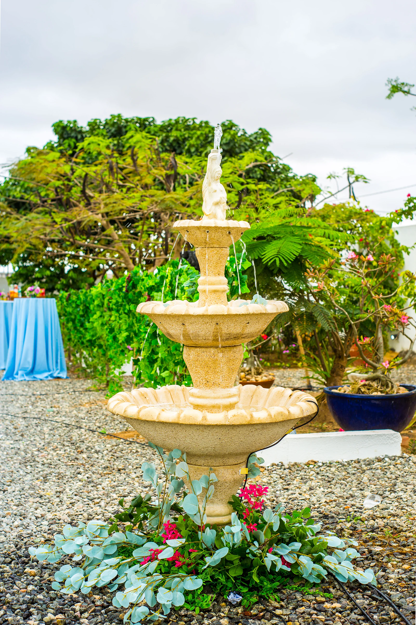 A three-tier stone fountain with eucalyptus and pink flowers at its base, on a gravel path.