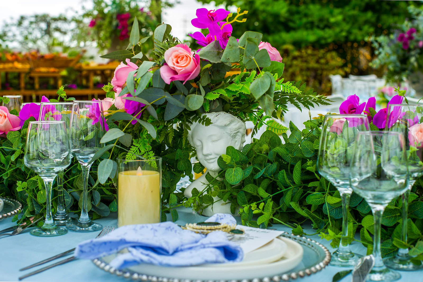 A centrepiece of pink roses, fuchsia orchids and foliage in a vase with a sculpted face, beside a candle in a glass holder.