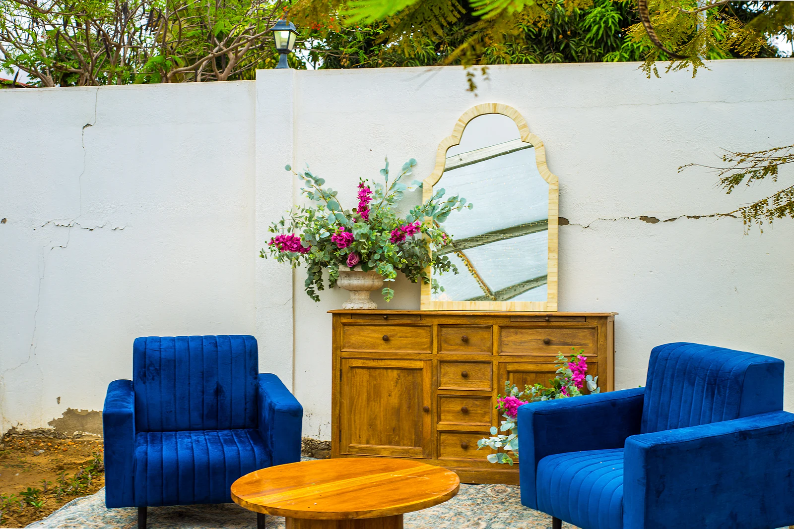 Two blue velvet armchairs, a low wooden table and a sideboard with an arched mirror and flowers, against a white wall.